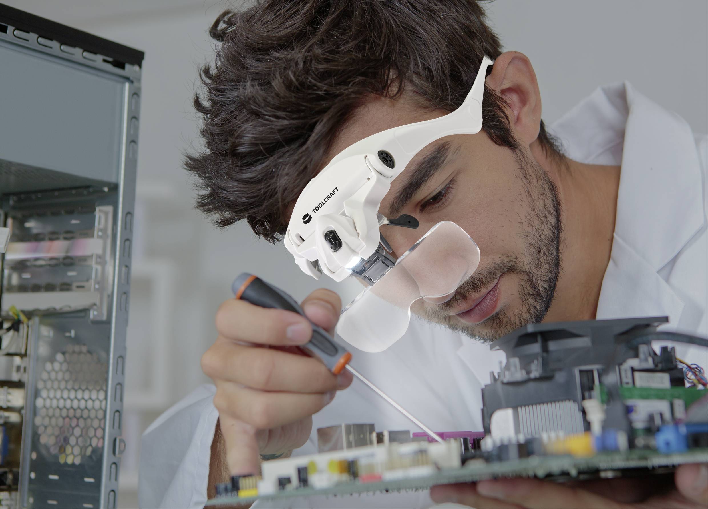 Technik v bílém plášti soustředěně pracuje na otevřené počítačové desce. Má nasazené zvětšovací brýle.