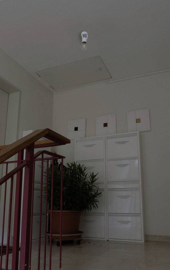 Kancelářská chodba s rostlinami, archiváři a schodištěm. Žárovka visí ze stropu. Stěny jsou natřeny bíle.