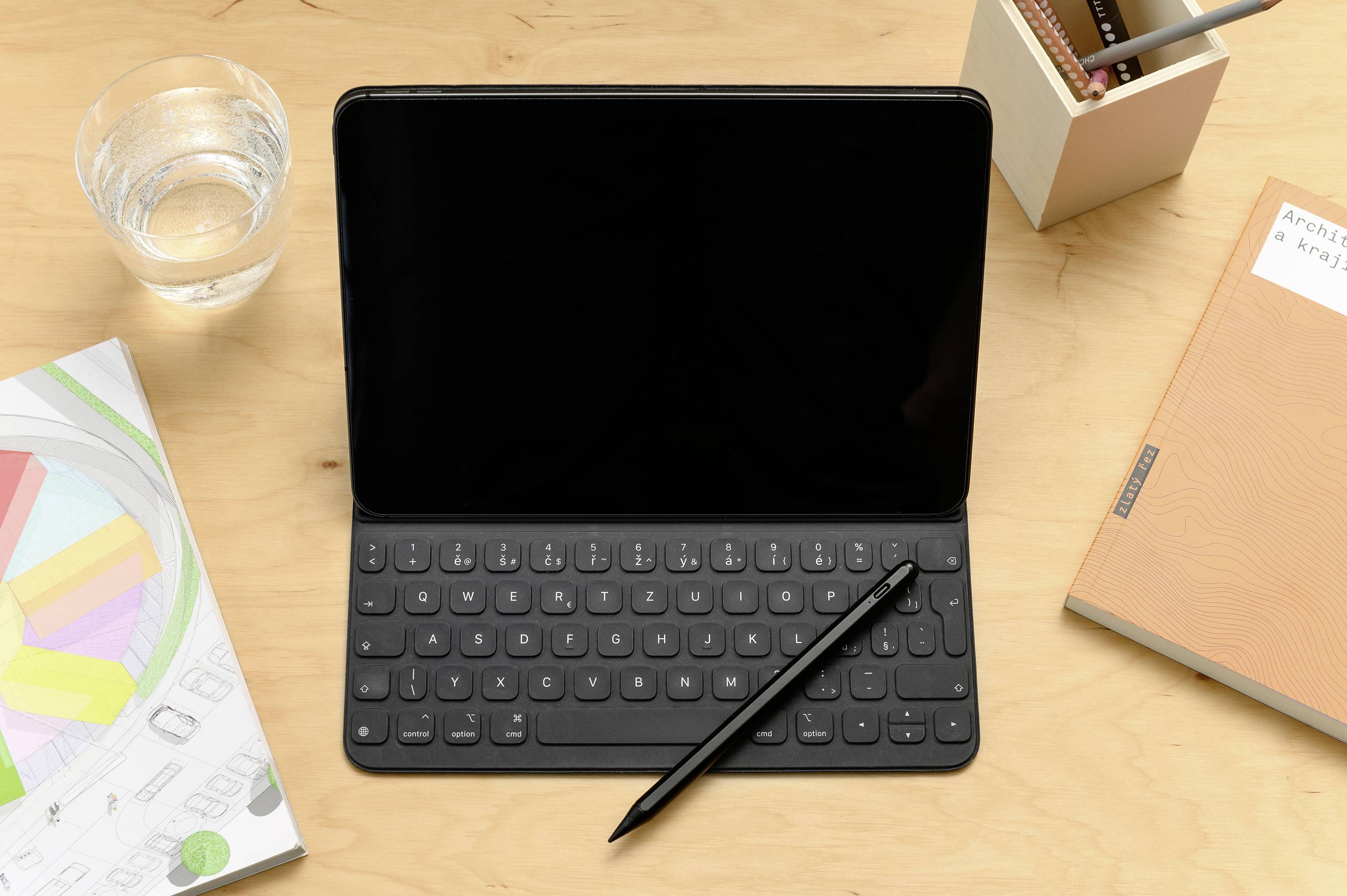Tablet s klávesnicí a perem na dřevěném stole, obklopený sklenicí vody, poznámkovým blokem a pouzdrem na tužky. Ideální pro práci nebo studium.