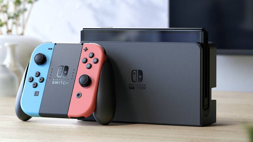 Nintendo Switch konzole umístěná na stole, připojená modrým a červeným ovladačem Joy-Con. V rozostřeném pozadí je vidět interiér.