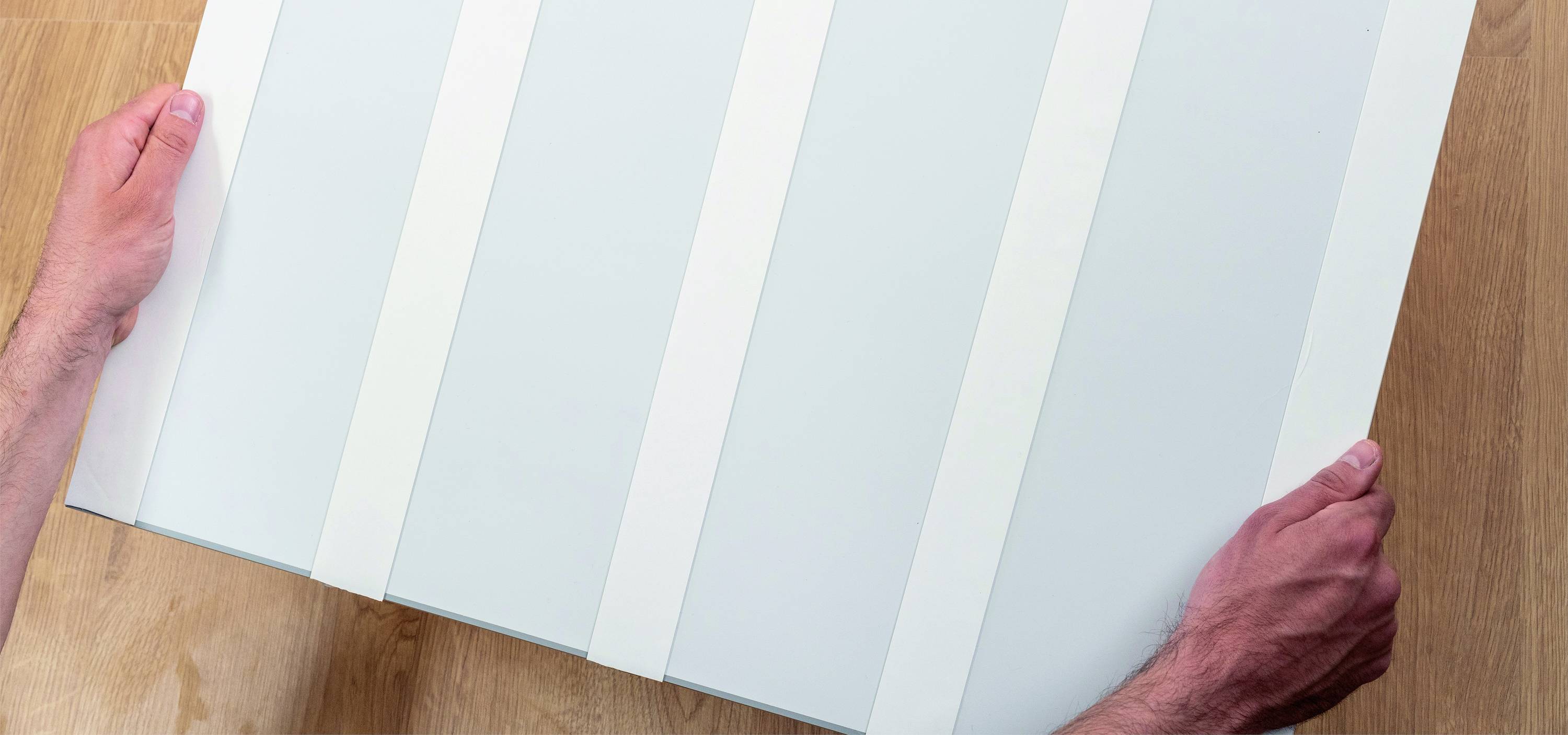 Dvě ruce drží velký, světlý panel s několika pravidelně rozmístěnými bílými pruhy, který leží na dřevěném stole.