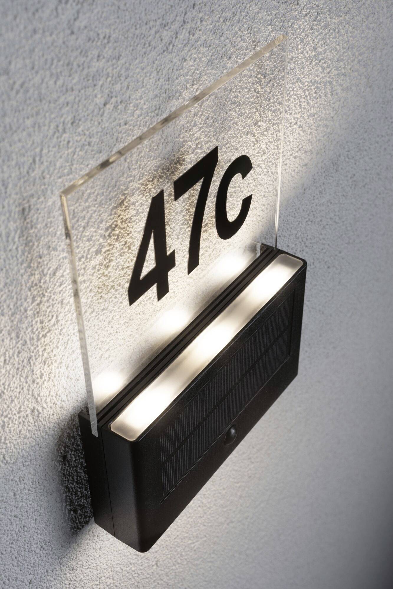 Osvětlená cedule s číslem domu '47c' na šedé zdi.