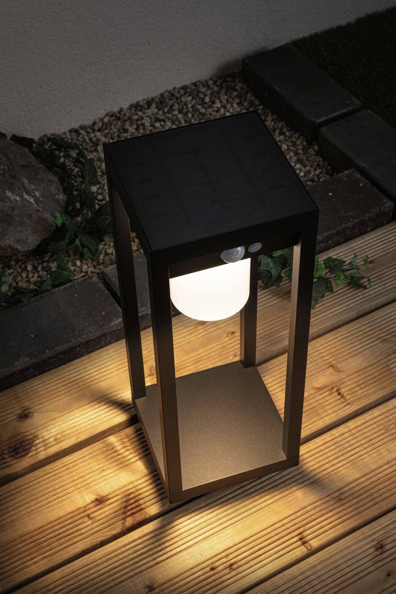 Solární zahradní lampa svítí na dřevěné terase. Má jednoduché, moderní designové prvky a poskytuje teplé světlo.
