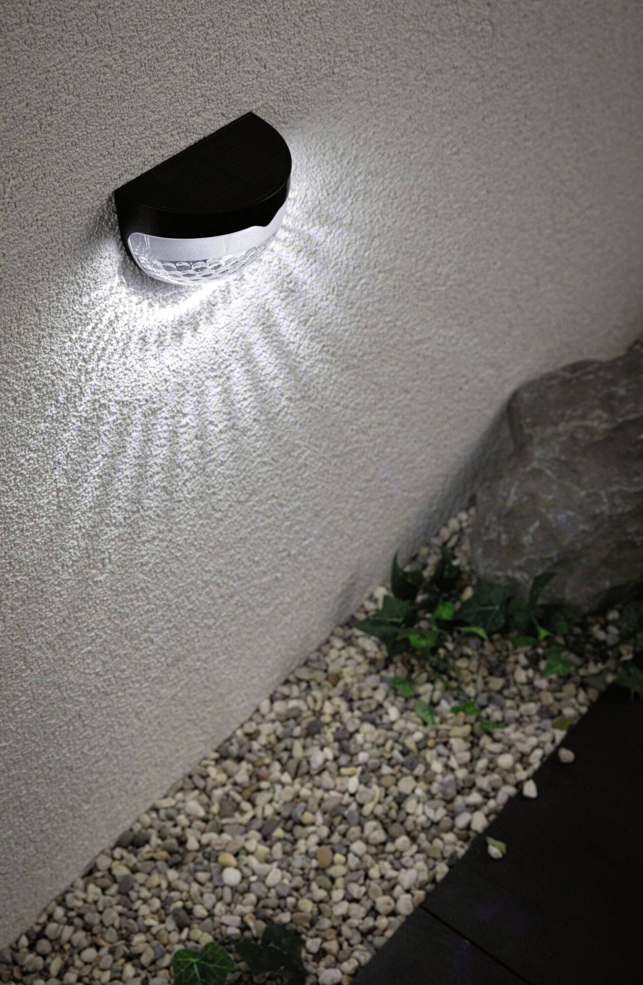 Nástěnná lampa osvětluje kamennou stěnu a rozptyluje světelné paprsky. Pod ní jsou viditelné oblázky a velký kámen.