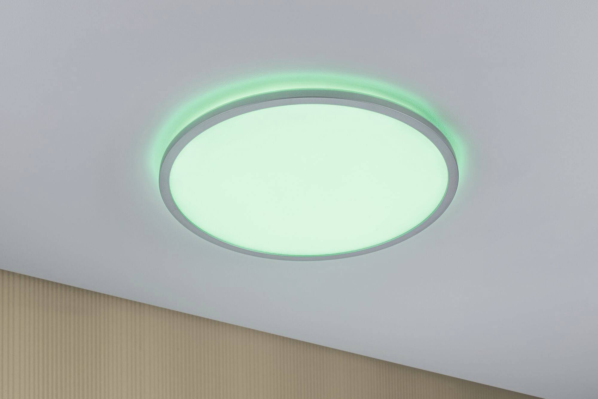 Kulatá stropní lampa se zelenavým světlem v místnosti, připevněná ke světlé stropní ploše. Osvětluje prostor rovnoměrně.