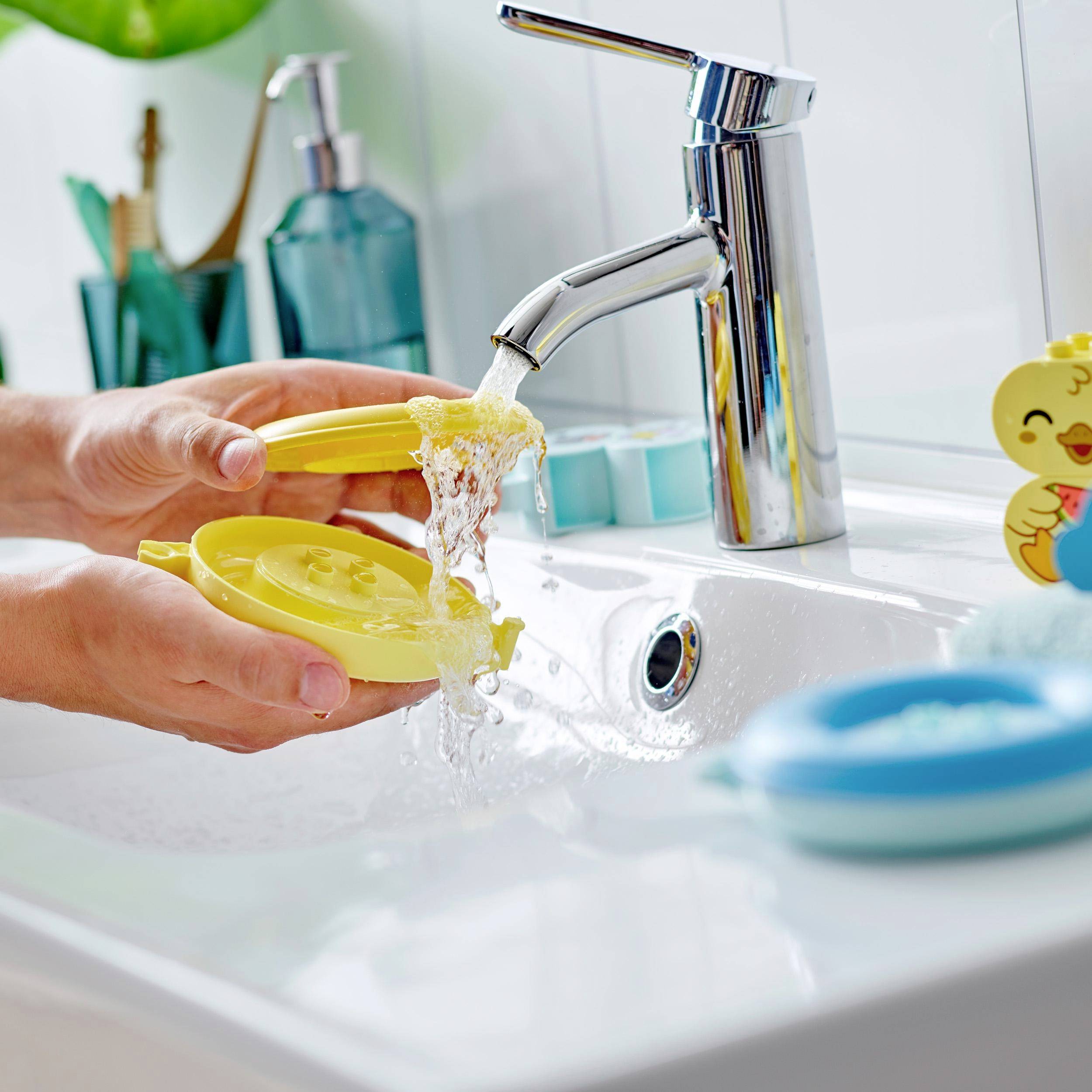 Člověk myje žlutý víko nádoby pod tekoucí vodou v umyvadle. Stranou stojí dávkovač mýdla a kachnička.