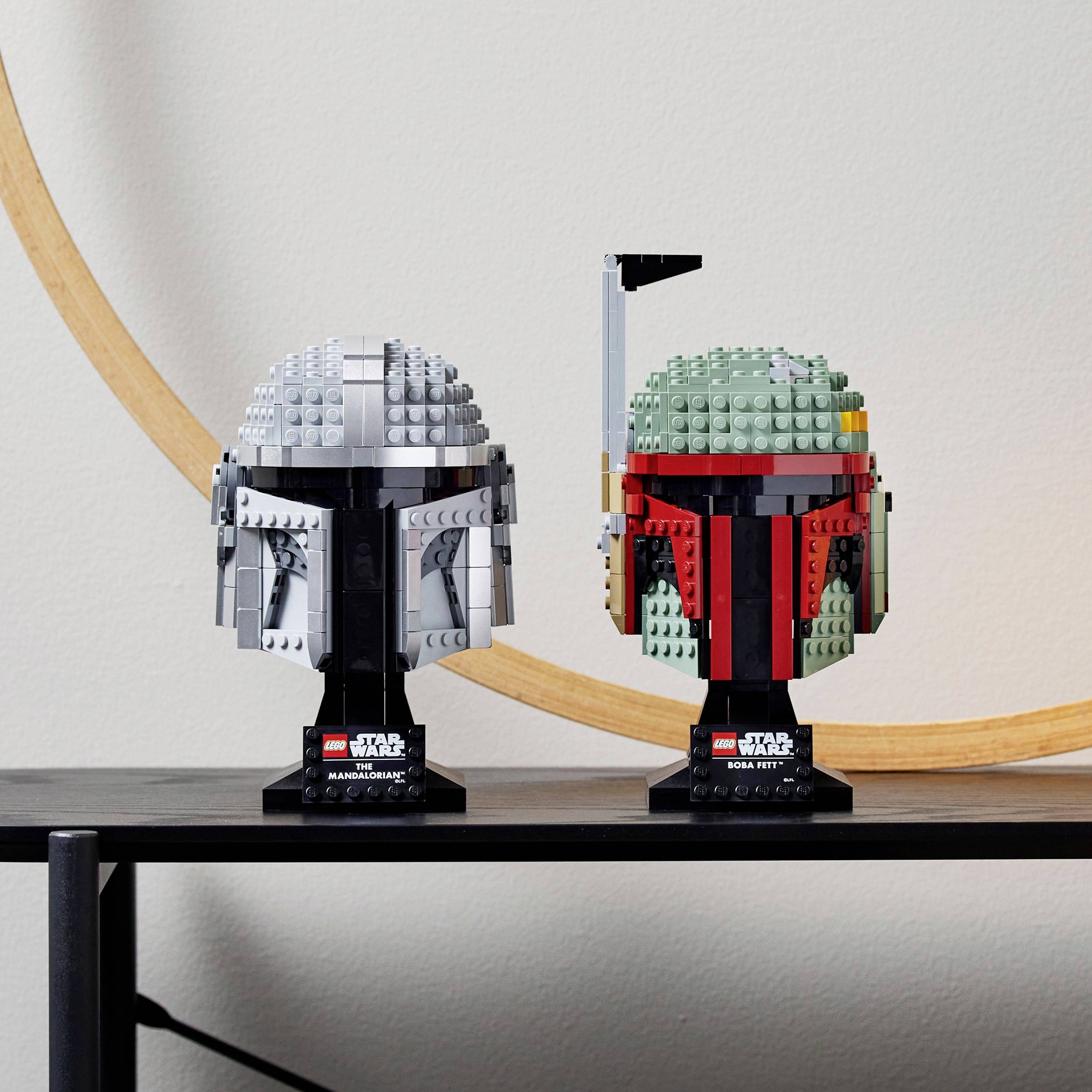 Dva modelové helmy LEGO z „Star Wars" na polici. Vlevo šedý helm, vpravo helm zeleno-červený. Široký oblouk v pozadí.