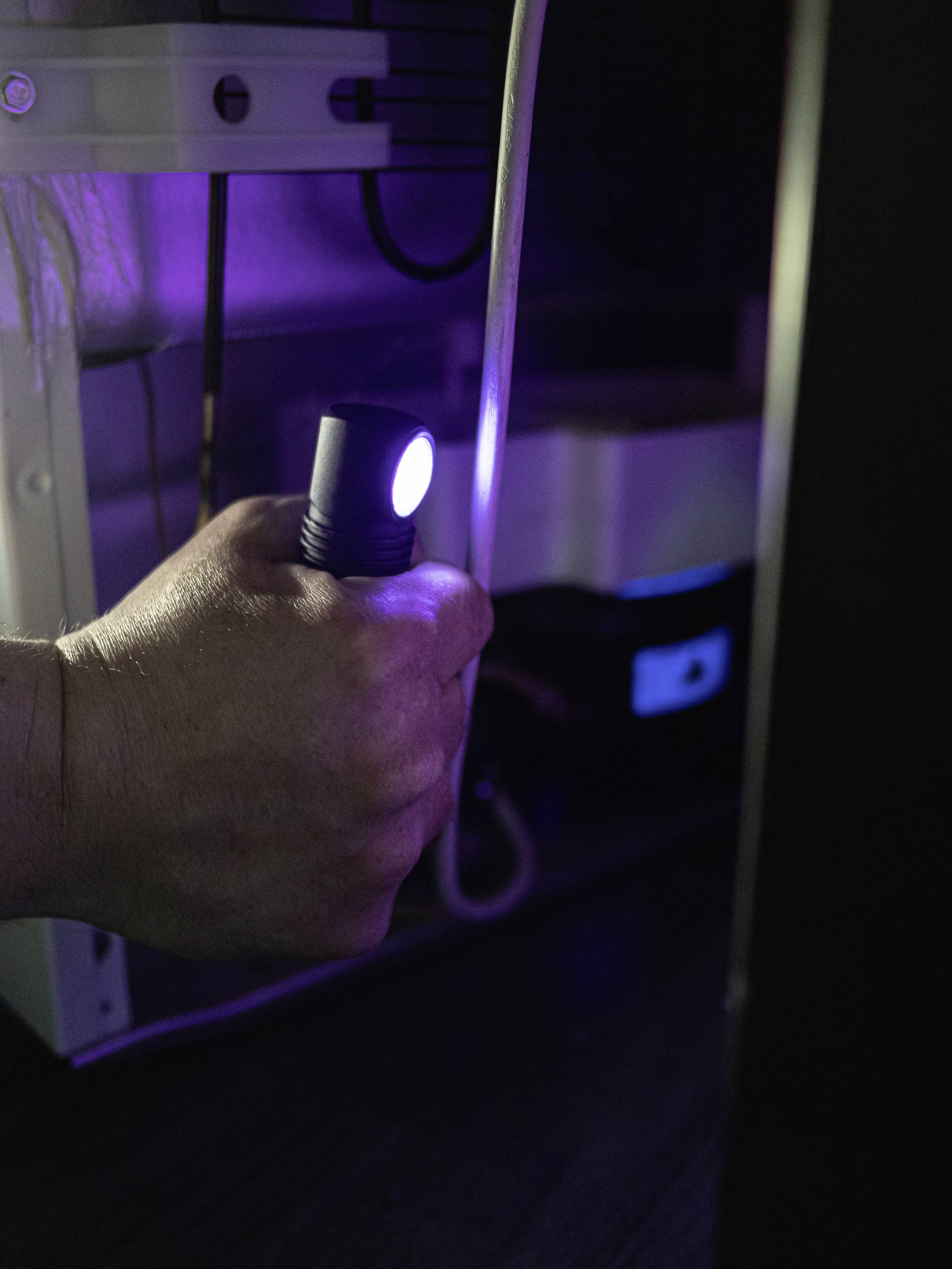 Ruka drží svítilnu a osvětluje kabely pod stolem. V pozadí jsou viditelné elektronické přístroje.