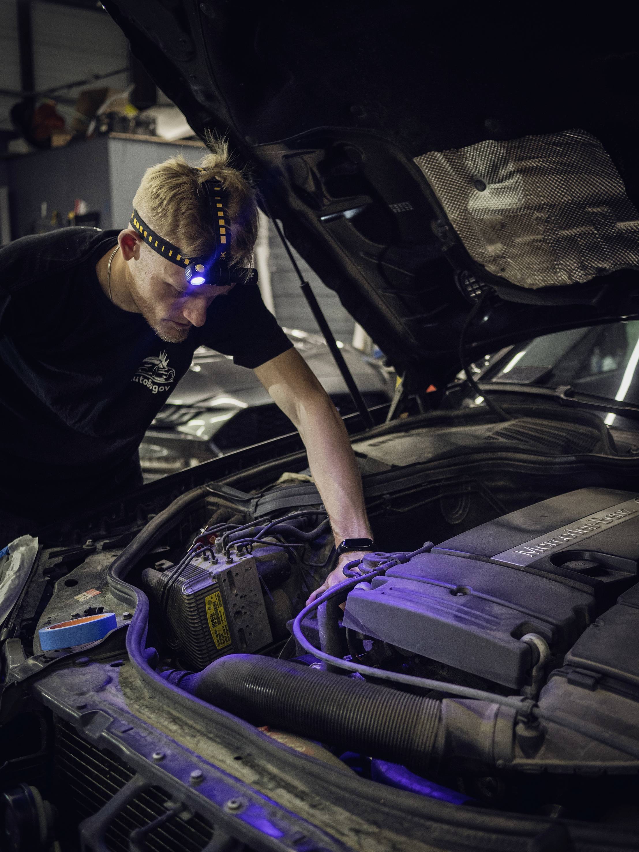 Mechanik s čelovkou kontroluje motor automobilu v autoservisu. Kapota je otevřená a on pracuje s plným soustředěním.