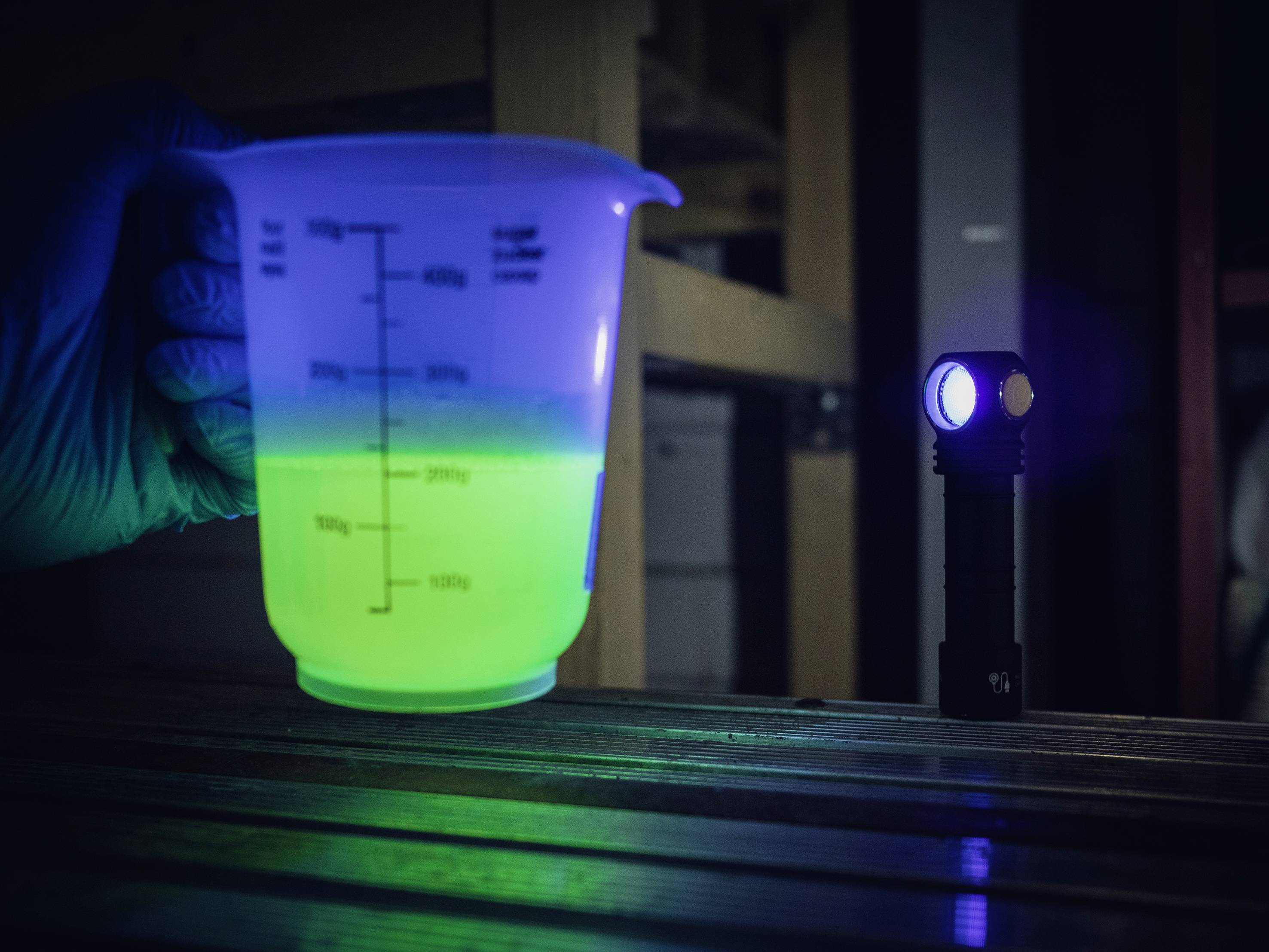 Jedna ruka v rukavici drží šálek s fluoreskující zelenou tekutinou, osvětlený malou baterkou.