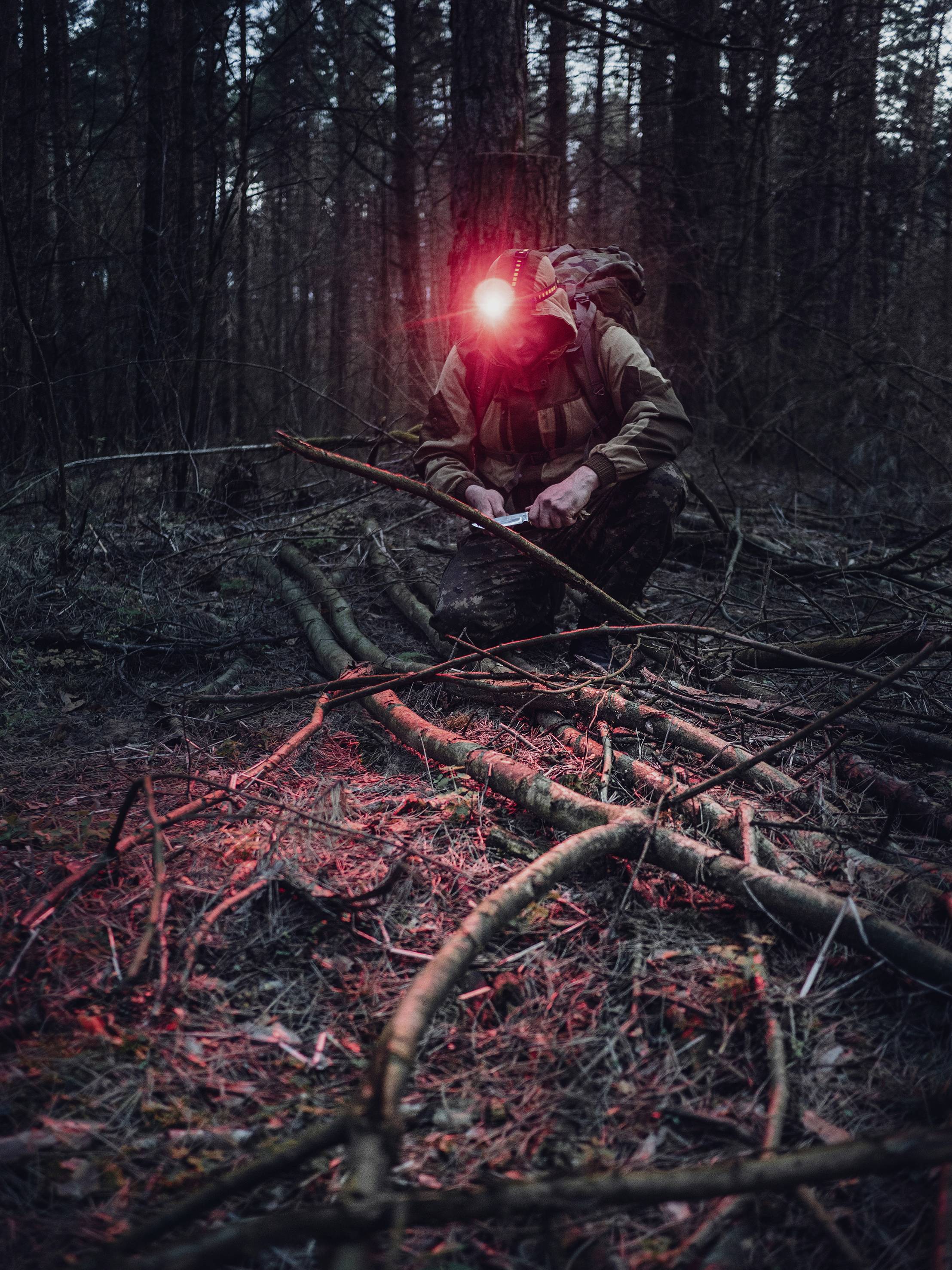 Osoba v tmavém lese, klečící nad nasbíranými větvemi. Má na zádech batoh a na čele svítící čelovou lampu, která osvětluje scénu.