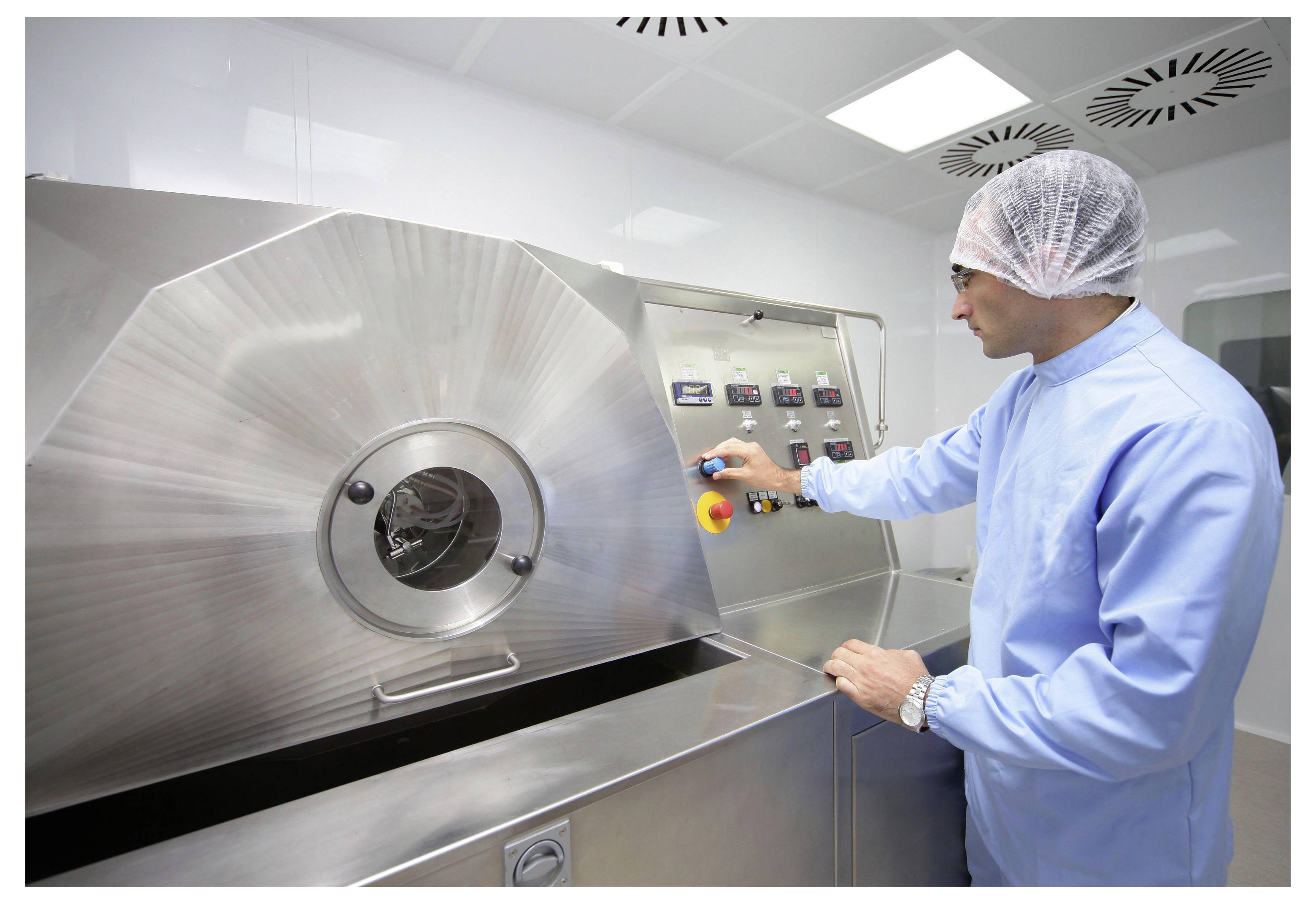 Technik v ochranném oblečení obsluhující velký průmyslový stroj z nerezové oceli v čisté místnosti nebo v oblasti farmaceutického zpracování.