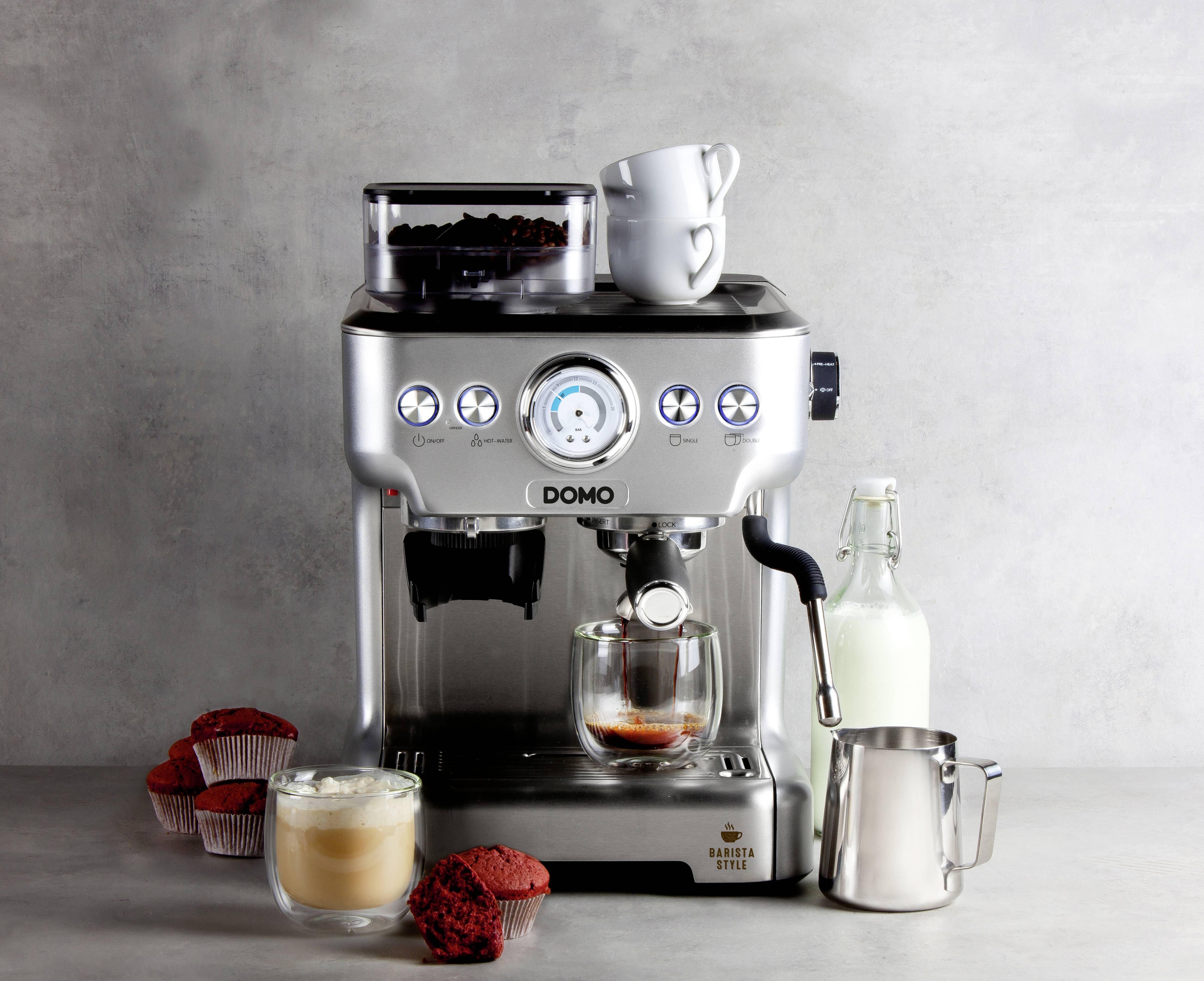 Espresso kávovar s napařující se kávou ve skle, obklopený muffiny, lahví mléka a konvičkou na mléko na šedém pozadí.