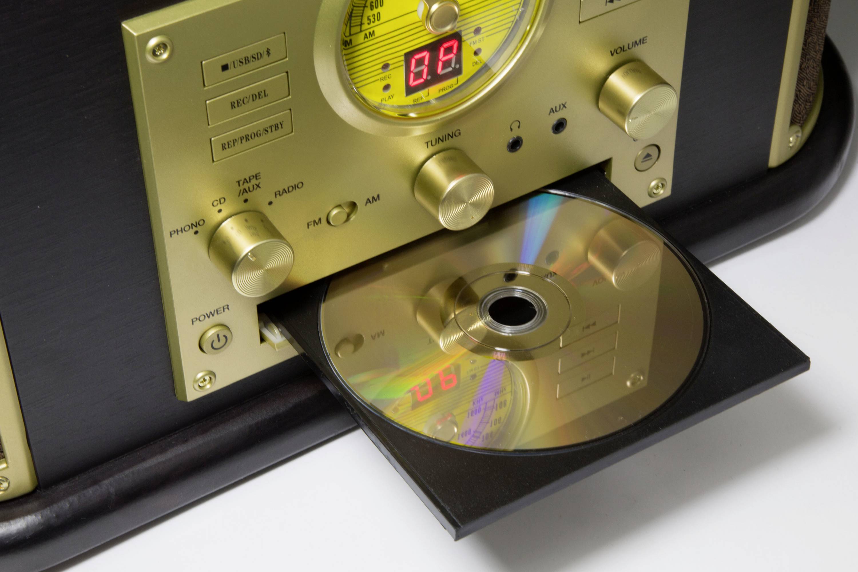 Retro rádio s otevřeným CD slotem, z něhož vyčnívá CD. Zlaté ovládací knoflíky a analogový displej na přední straně.
