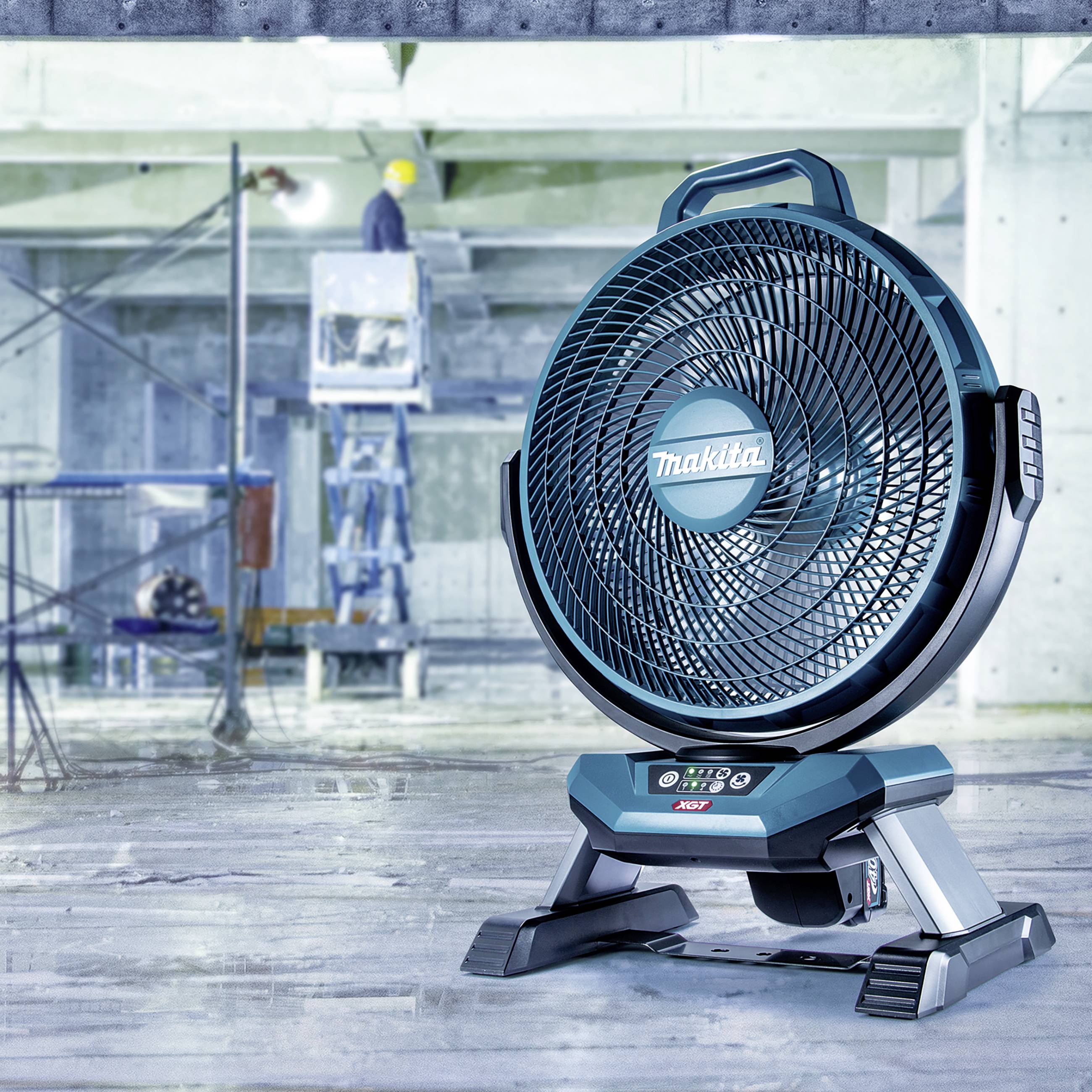 Modrý ventilátor stojí v nedokončené budově. V pozadí pracuje osoba v ochranné přilbě.