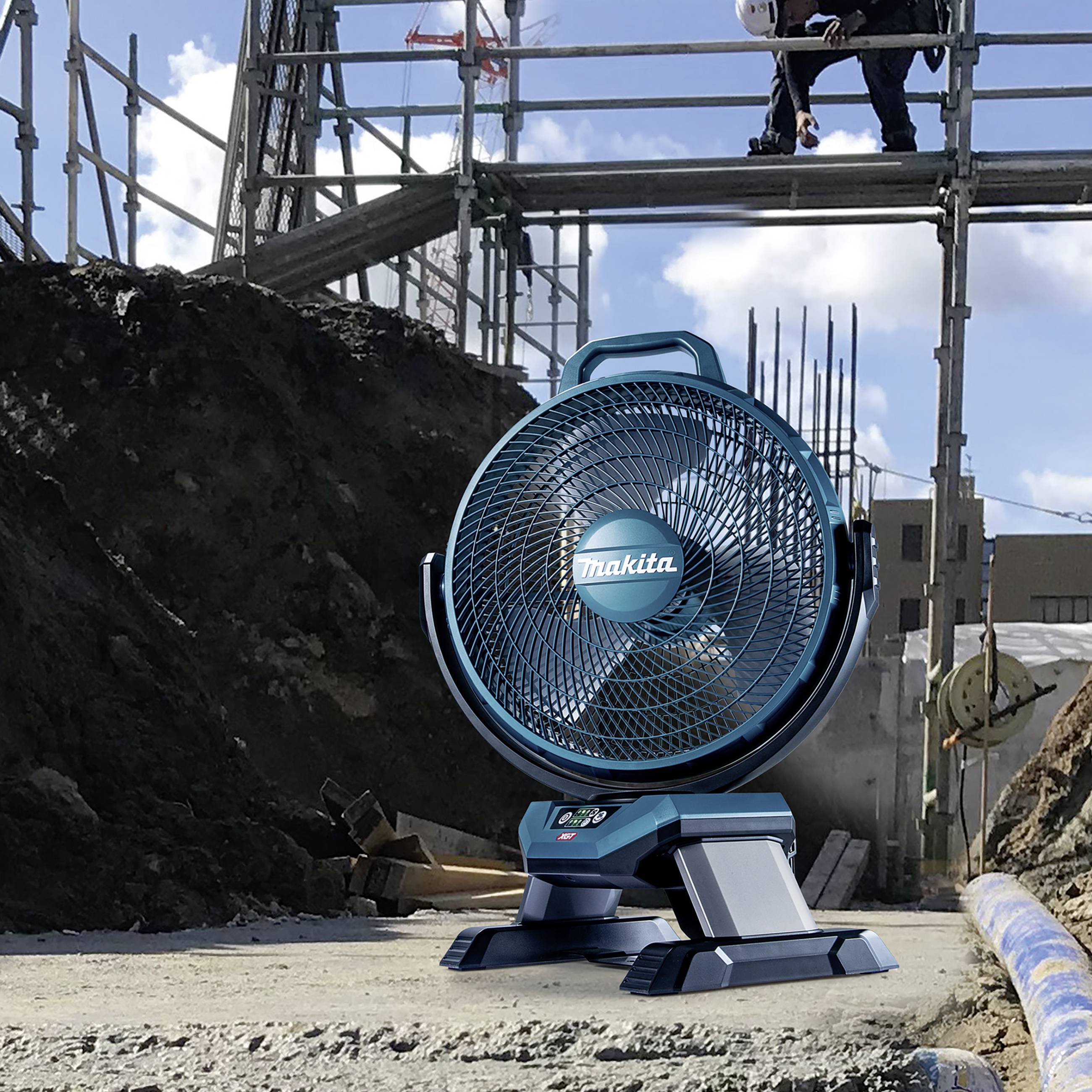 Stavební ventilátor stojí na zemi v popředí. V pozadí pracují lidé na lešení nad zemní jámou.