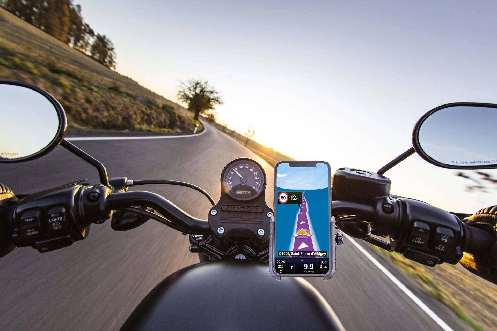 Motocykl jede při západu slunce po zatáčkovité silnici. Mobilní telefon s navigační aplikací v popředí ukazuje trasu do Lublaně.