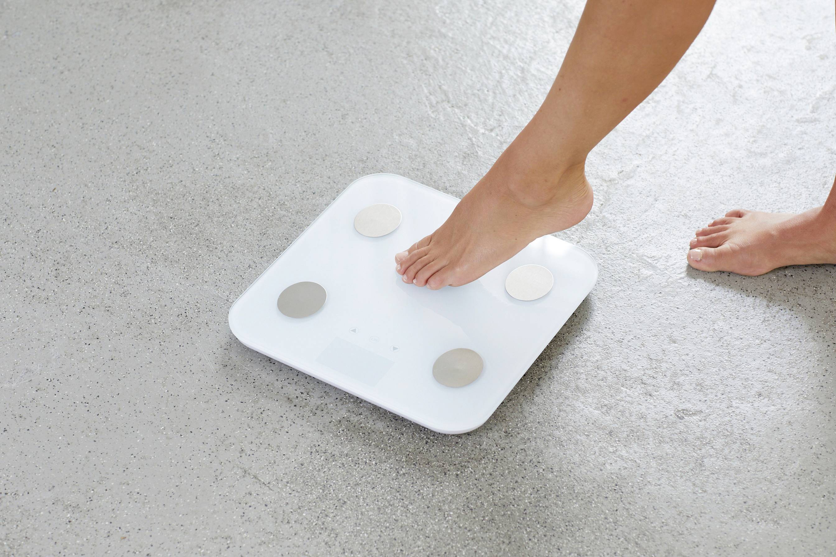 Noha se přibližuje bílé osobní váze na šedé podlaze. Váha má čtyři kovové senzory pro měření hmotnosti a tělesných parametrů.