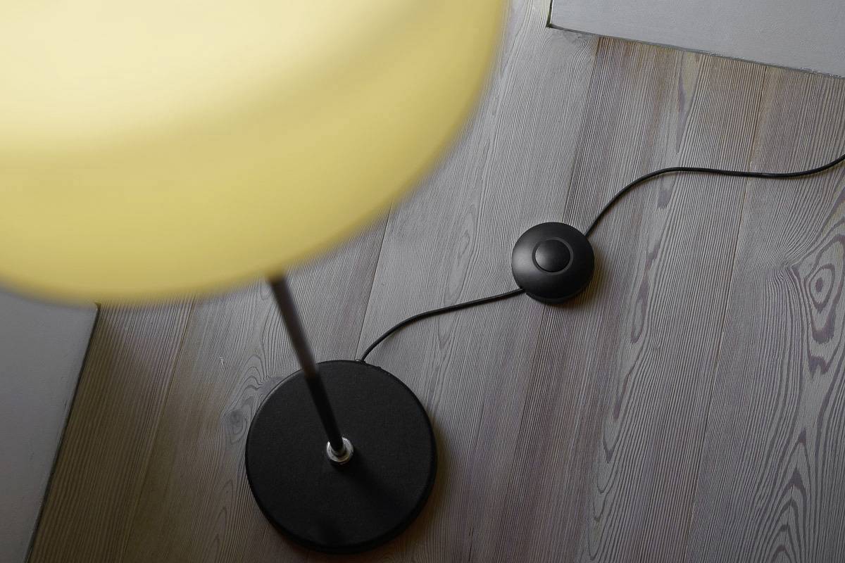 Detailní záběr stojací lampy se žlutým stínítkem, vedle níž leží černý nožní vypínač na dřevěné podlaze.