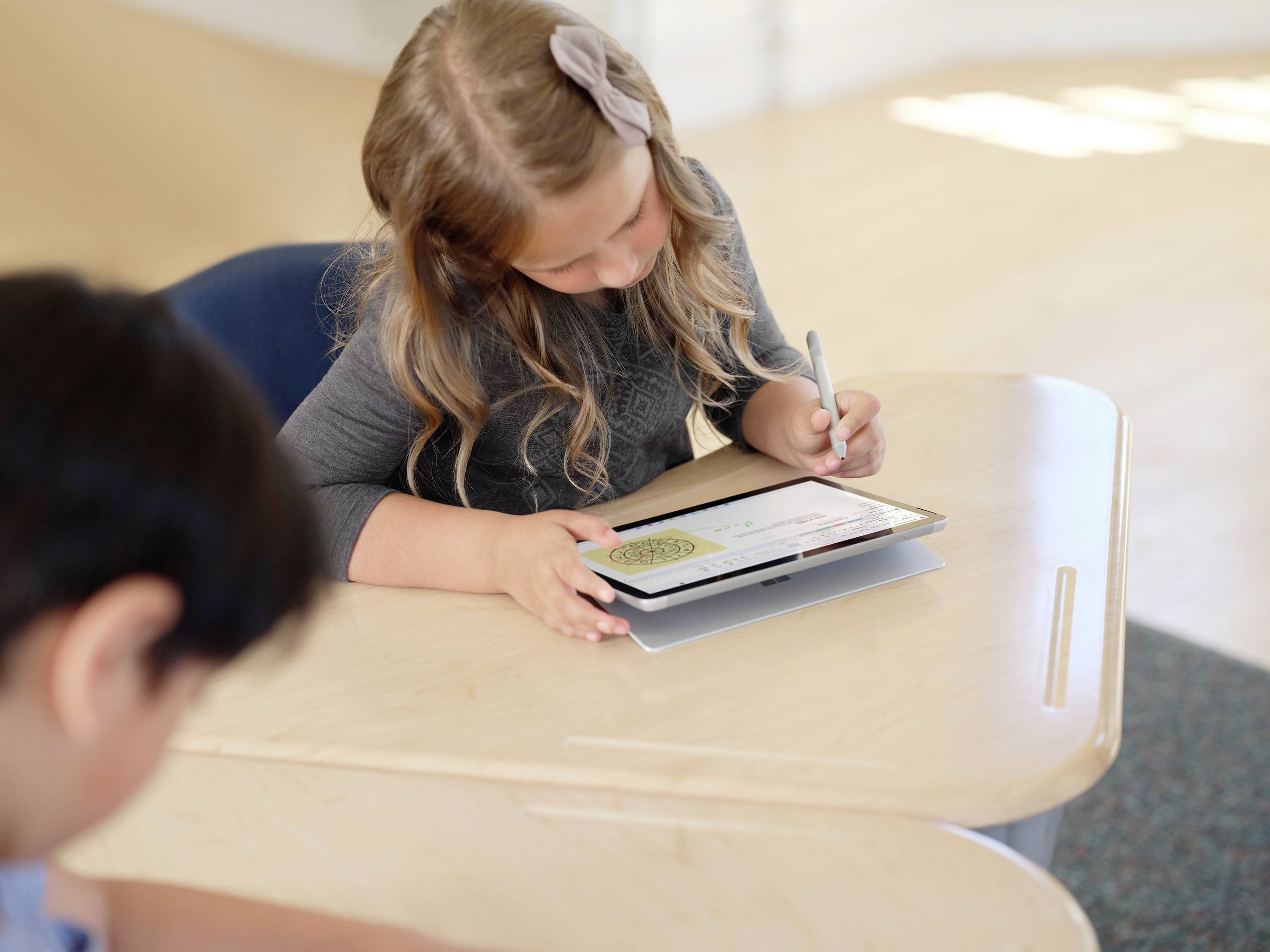 Dívka sedí u stolu a soustředěně pracuje s tabletem a perem. Další dítě je v popředí.