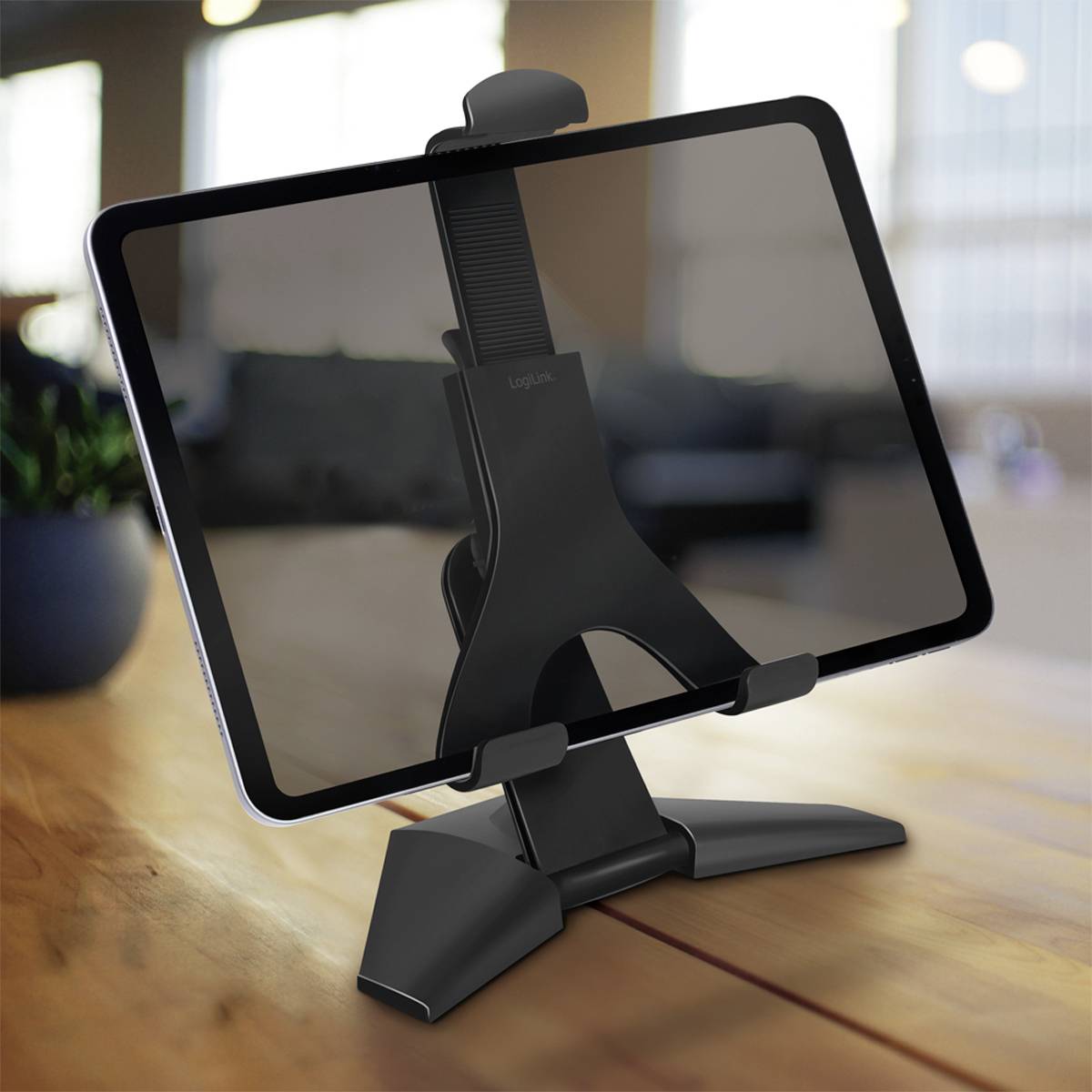 Tablet leží na černém stojanu na dřevěném stole v moderní místnosti. V pozadí jsou rozmazané kancelářské prostory.