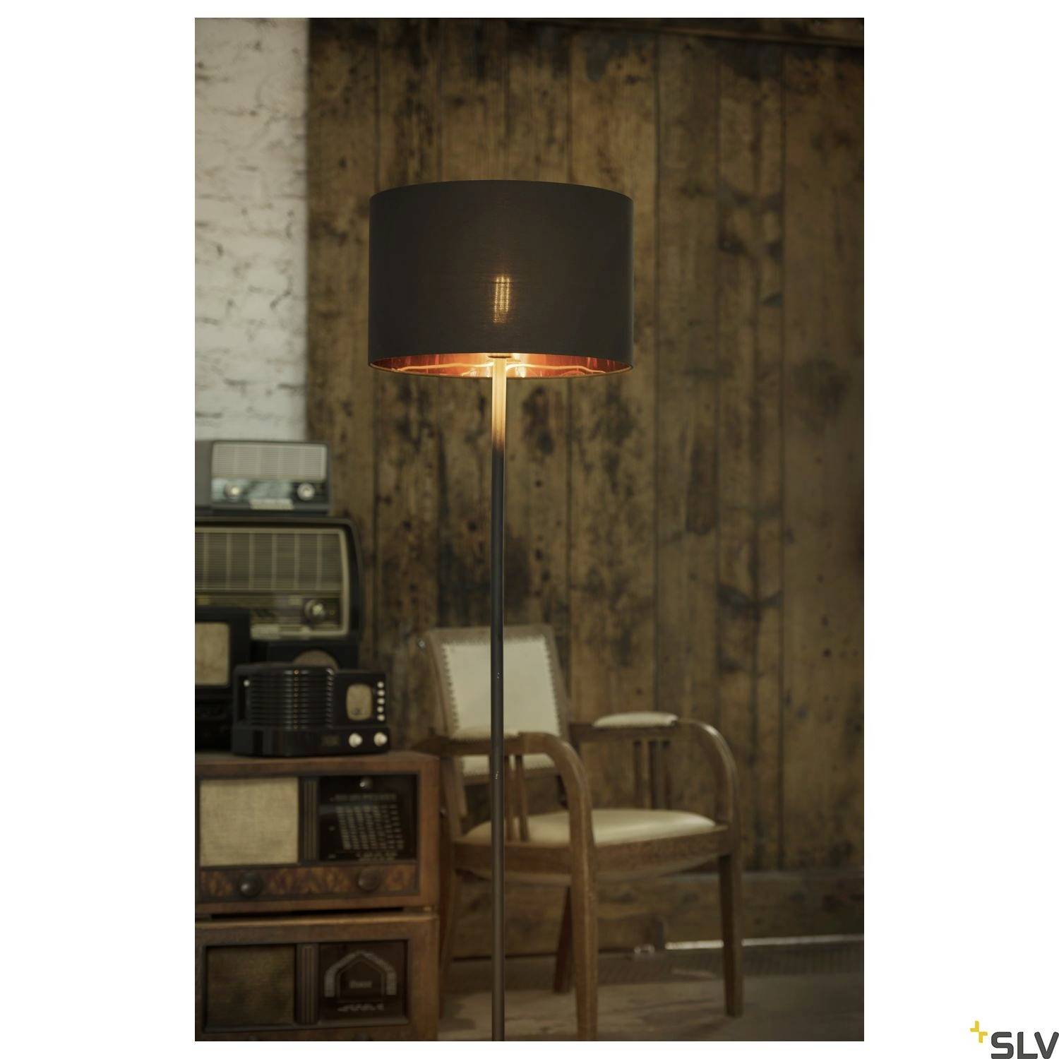 Stojací lampa s černým stínítkem svítí v místnosti s dřevěnými stěnami a retro nábytkem. Vintage rádia jsou umístěná na poličce v pozadí.
