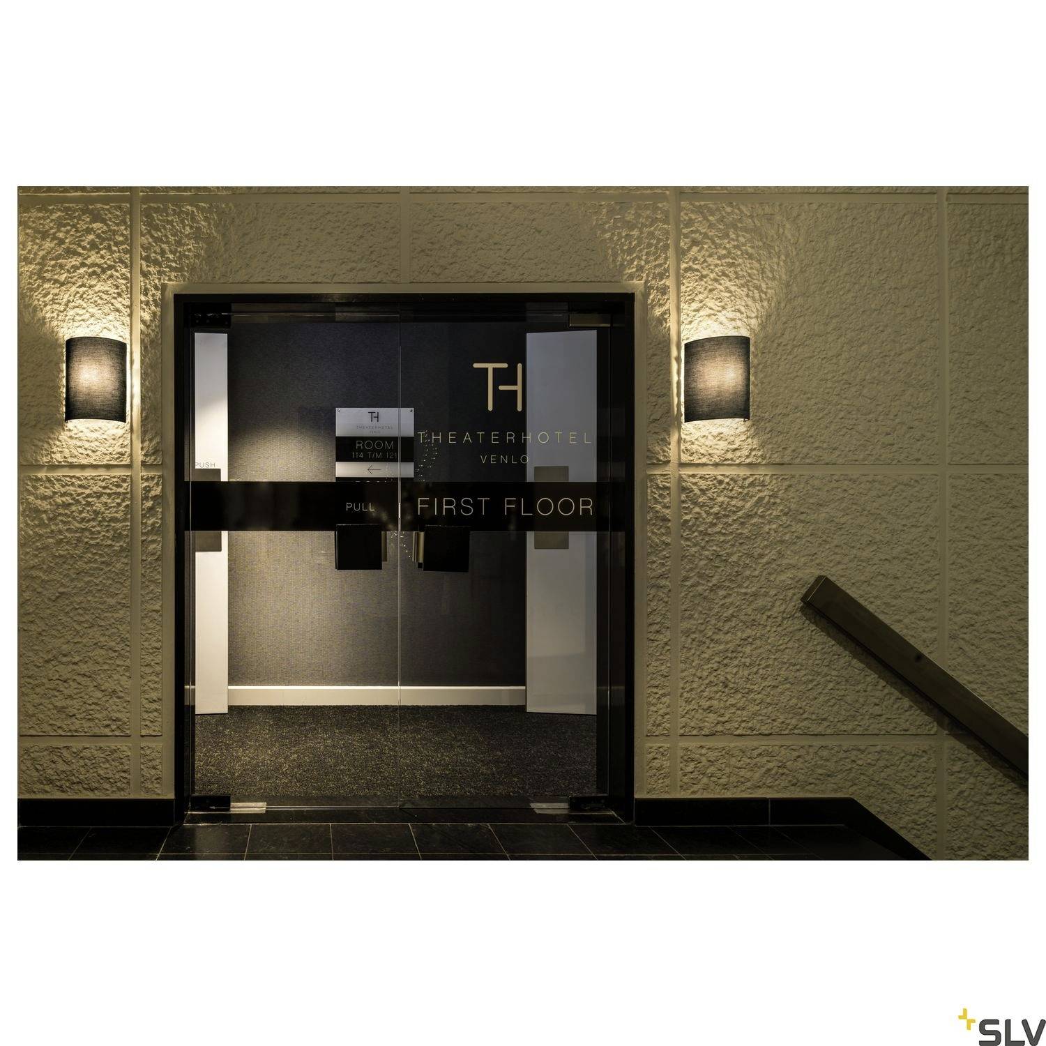 Obrázek zobrazuje vstup do hotelu se dvěma nástěnnými lampami osvětlujícími kovové dvoukřídlé dveře. Na dveřích je nápis 'PRVNÍ PATRO'.