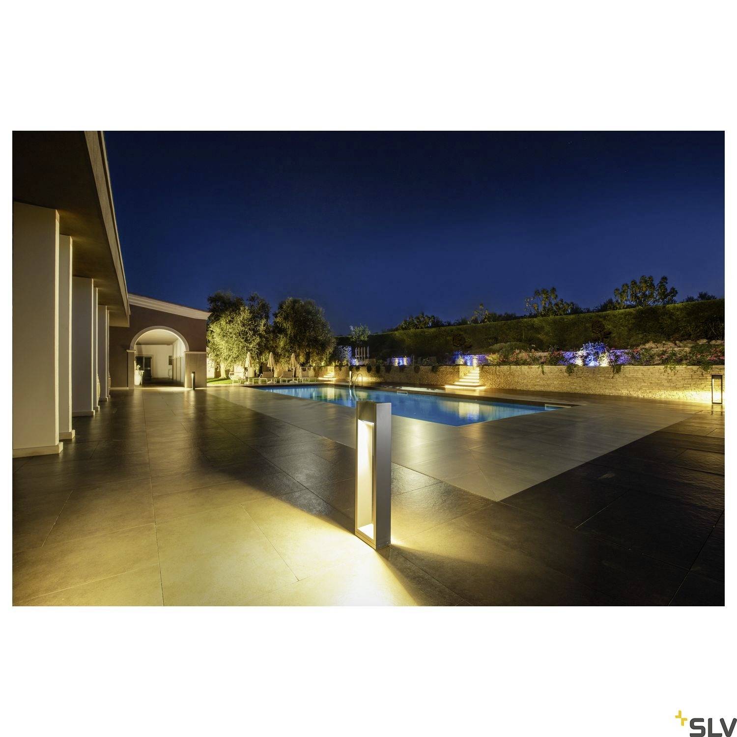 Noční scéna dobře osvětleného venkovního bazénu vedle moderní budovy. Stromy a rostliny obklopují prostor bazénu.