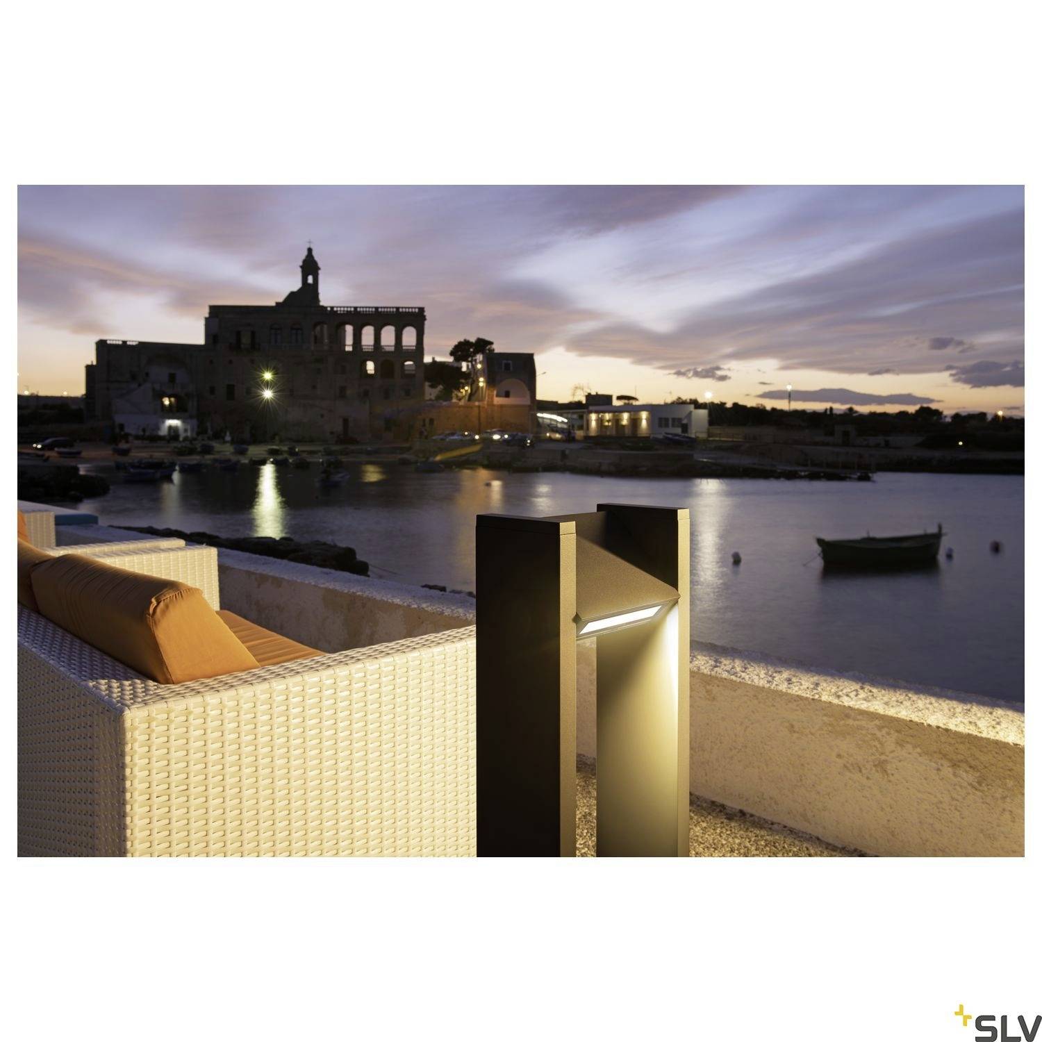 Večerní atmosféra u vodní hladiny s osvětlenou cestou a sedačkou v popředí, historickými budovami a klidnou oblohou v pozadí.