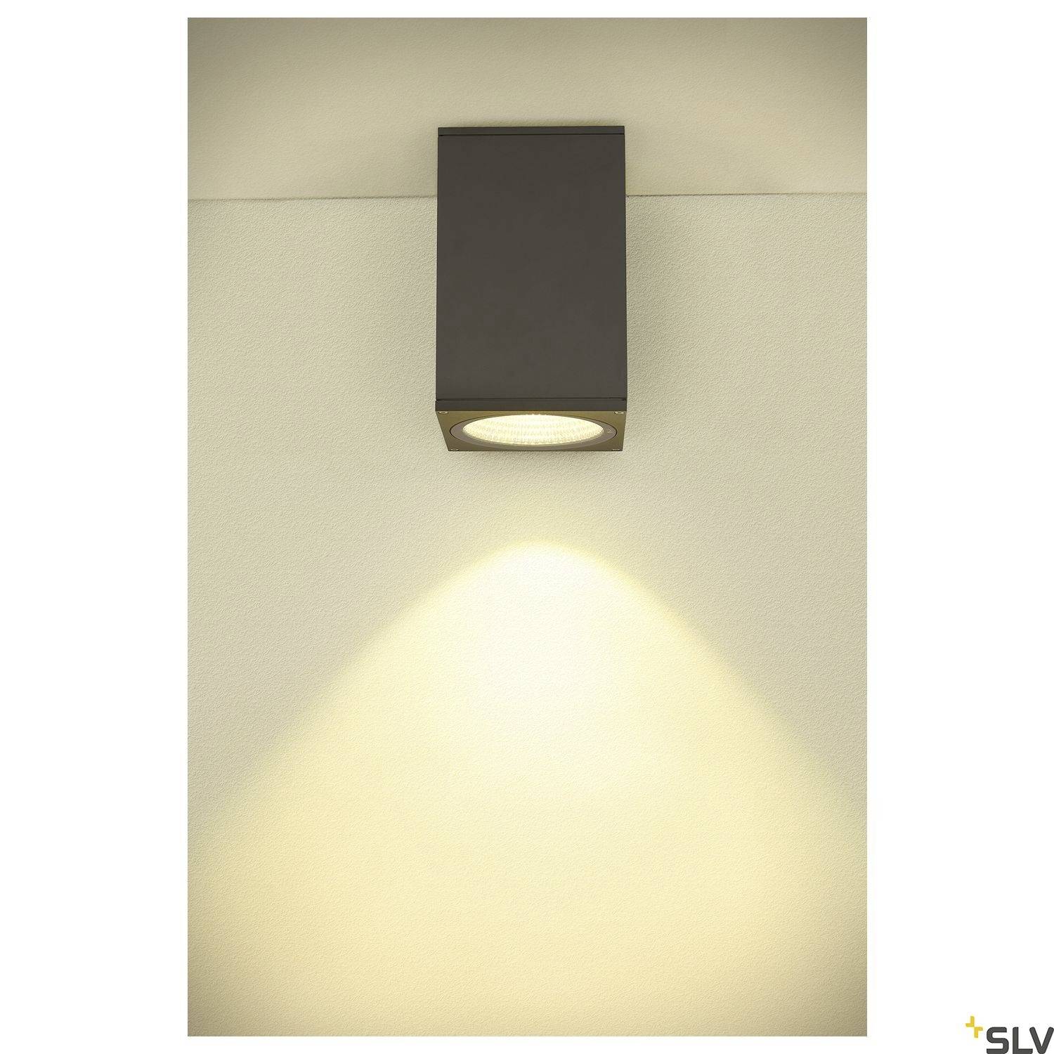 Nástěnné svítidlo, které osvětluje béžovou stěnu směrem dolů a vytváří jemný světelný vzor. Obdélníkový, moderní design v tmavě šedé barvě.