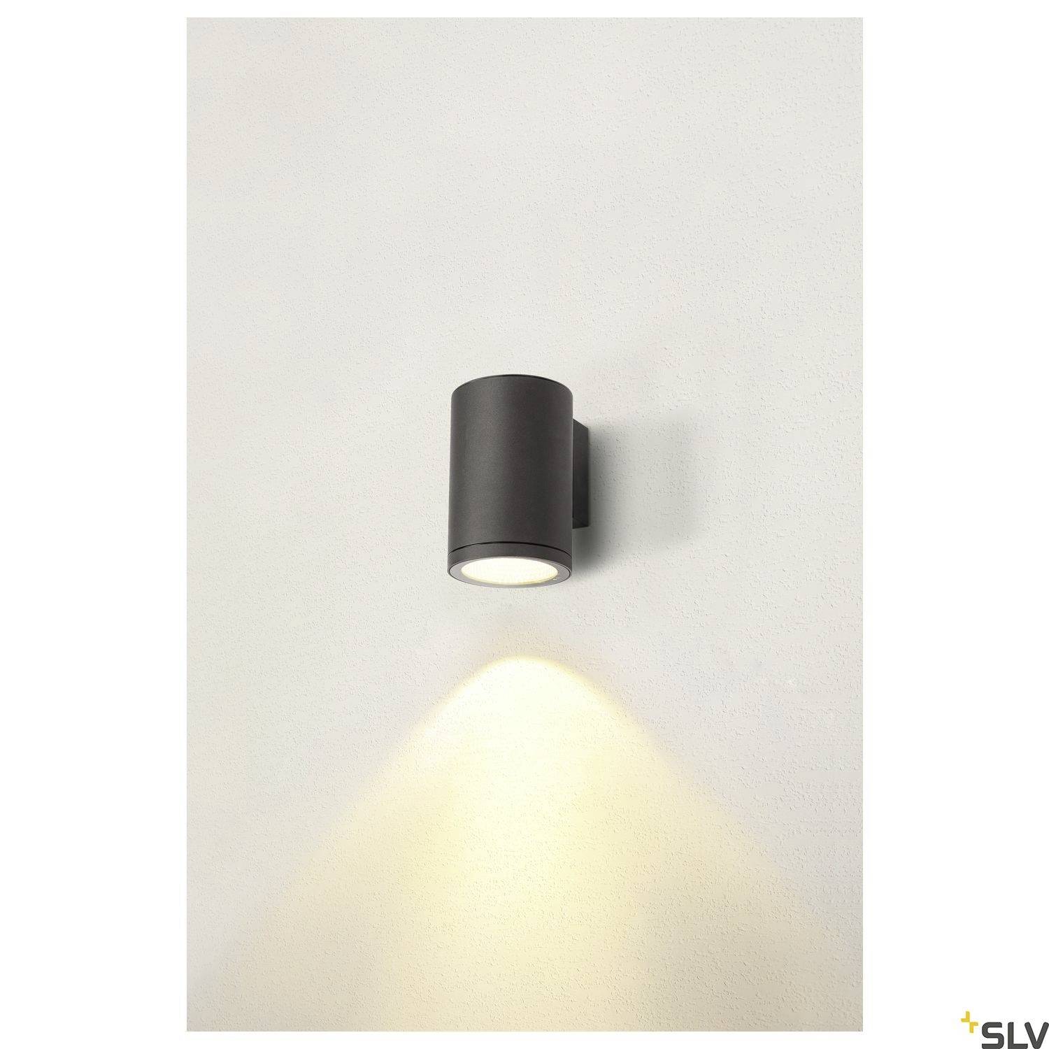 Černé nástěnné svítidlo, které svítí dolů, osvětluje prostor pod ním. Stěna kolem lampy je světle šedá.