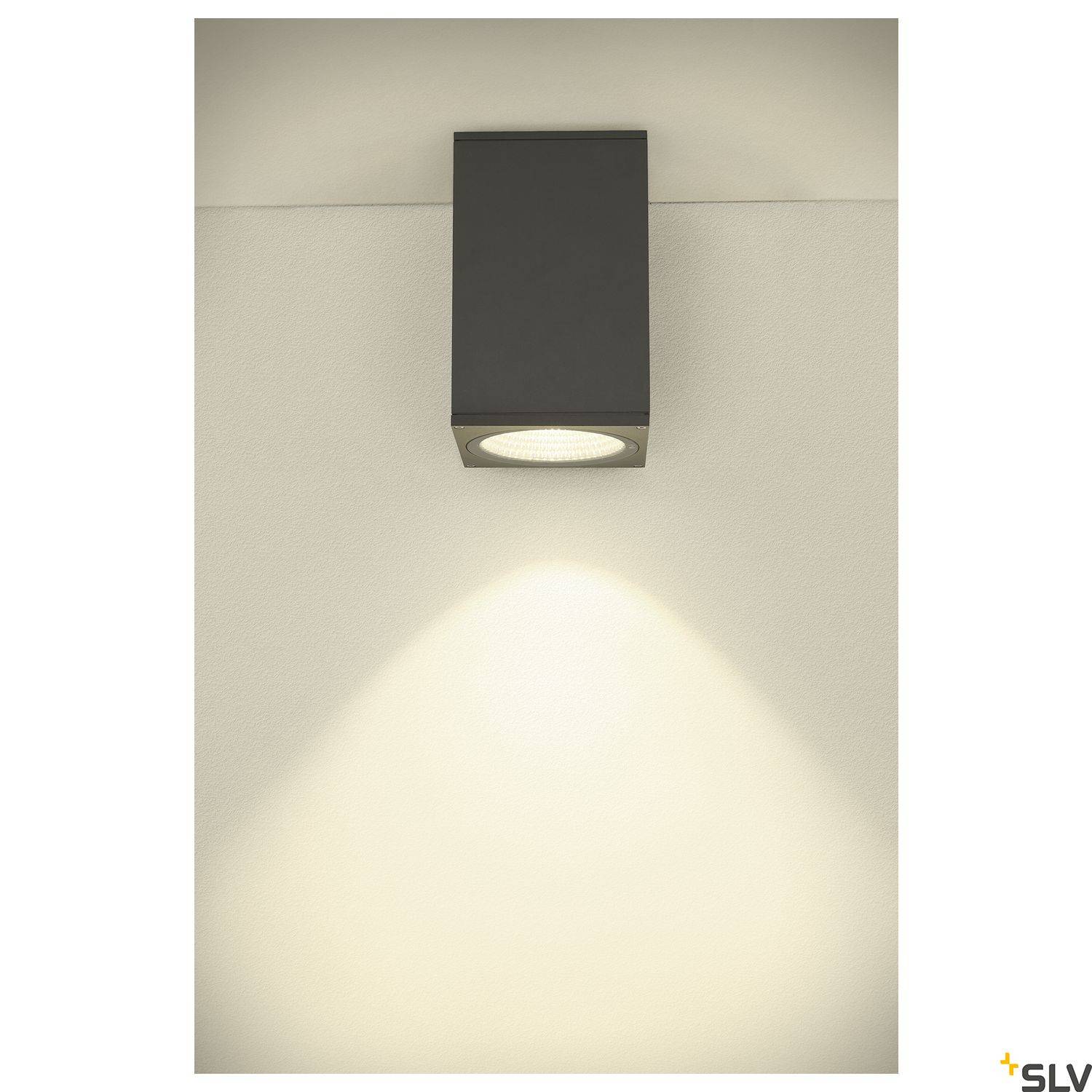 Nástěnná lampa s čtvercovým tělem vrhá kuželovitě tvarované světlo na stěnu.