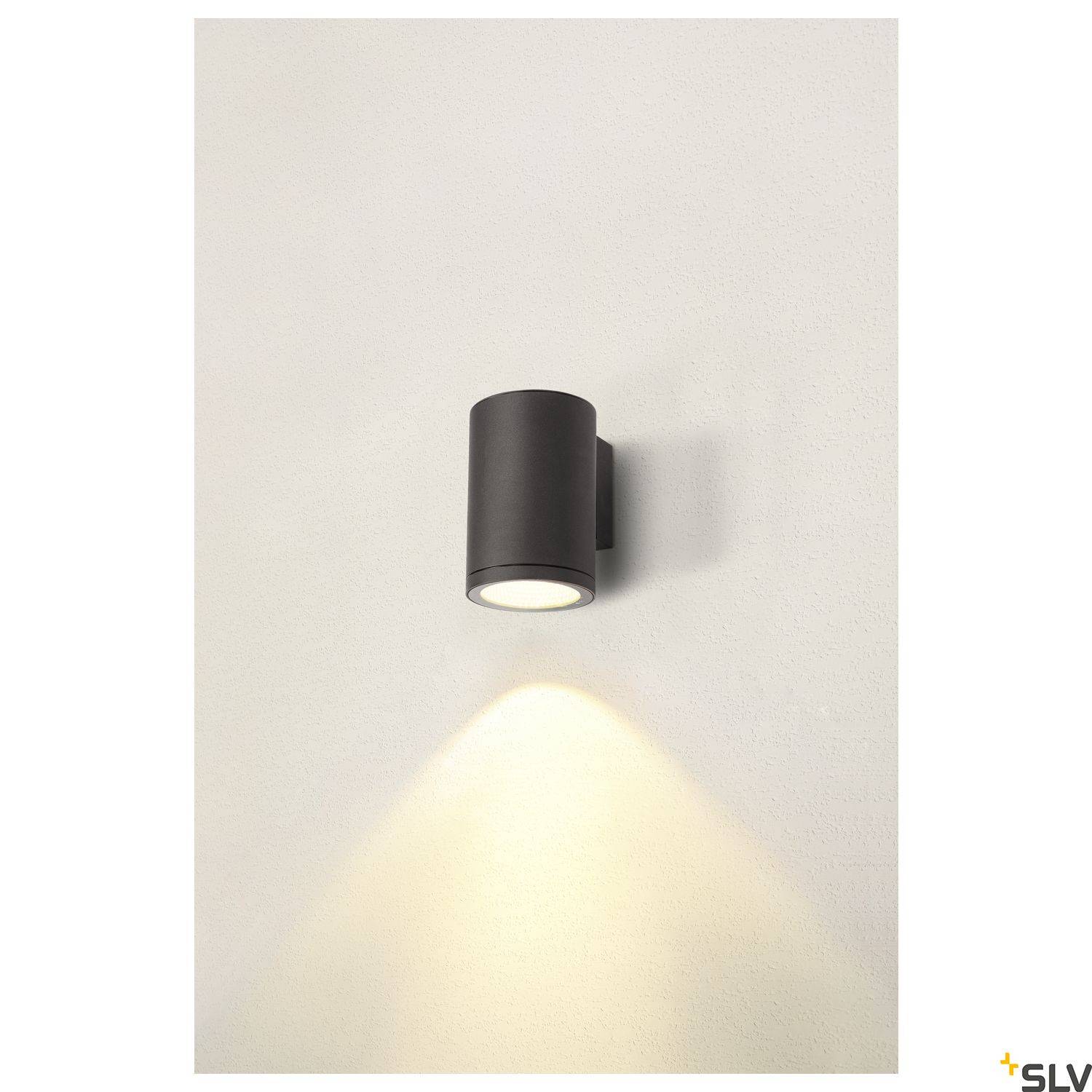 Černá válcovitá nástěnná lampa vrhá teplé světlo na bílou stěnu.