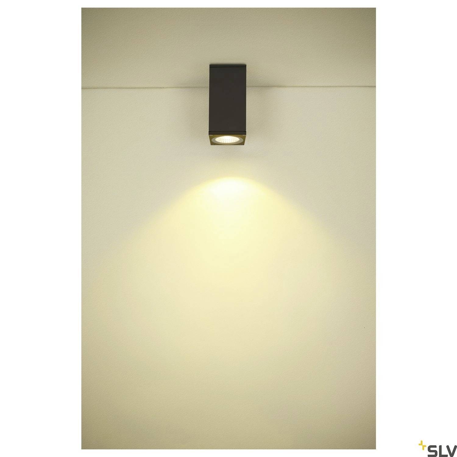 Obdélníková nástěnná lampa vyzařuje teplé světlo dolů. Stěna má světlé, neutrální pozadí.