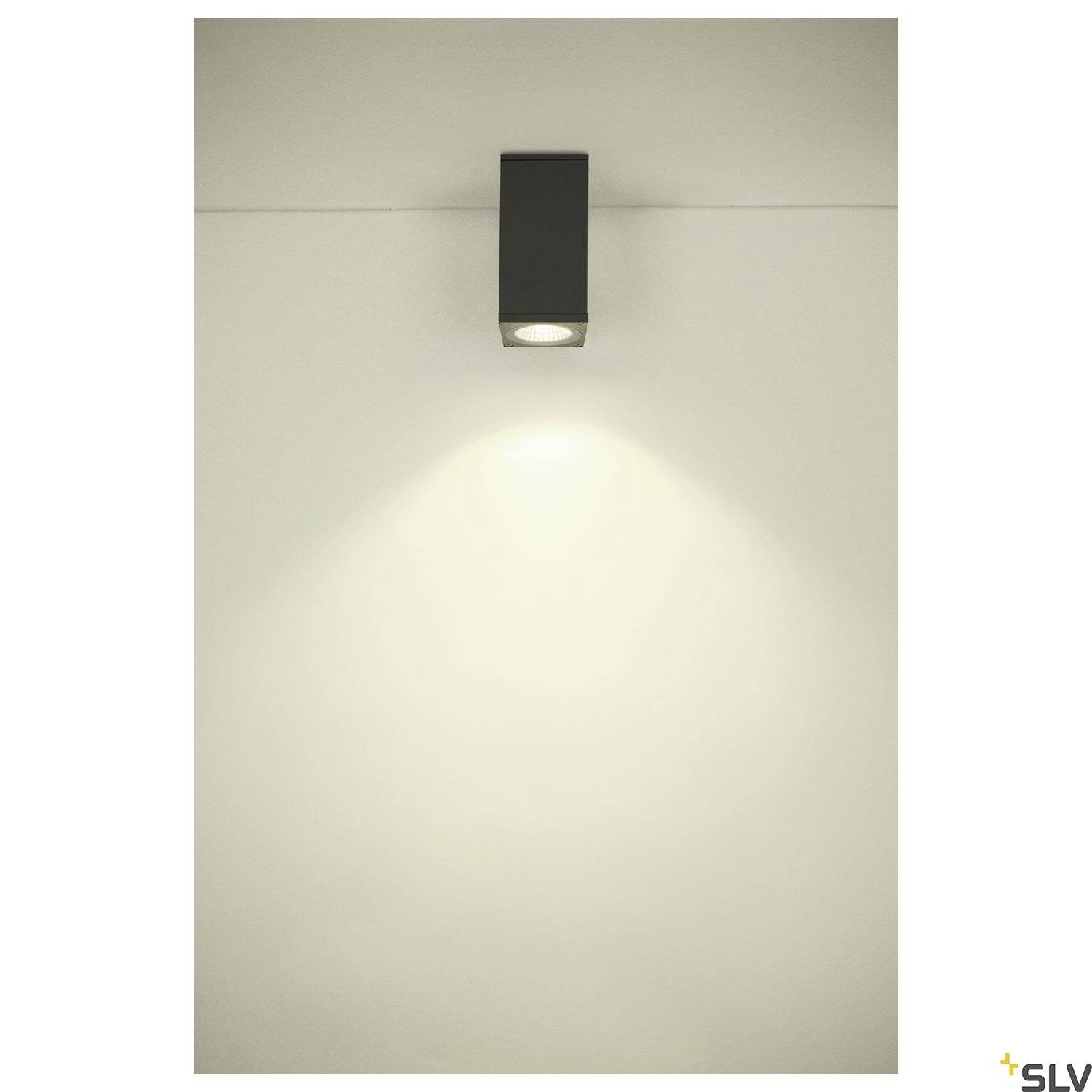 Minimalistická, obdélníková nástěnná lampa s černým tělem vrhá teplé světlo dolů na bílou stěnu.