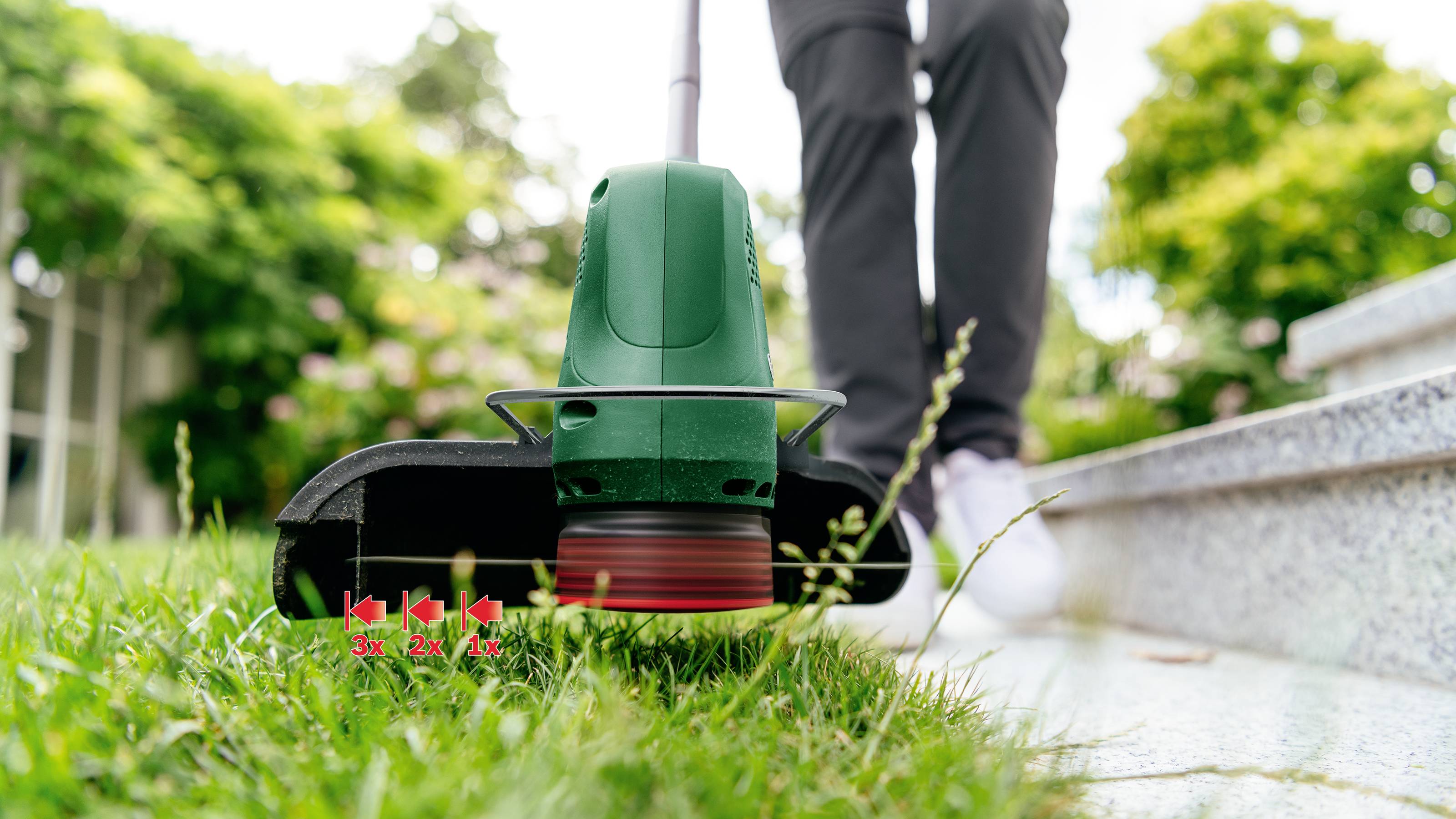 Osoba seká trávu podél chodníku zeleným travním křovinořezem. V pozadí jsou vidět stromy a keře.