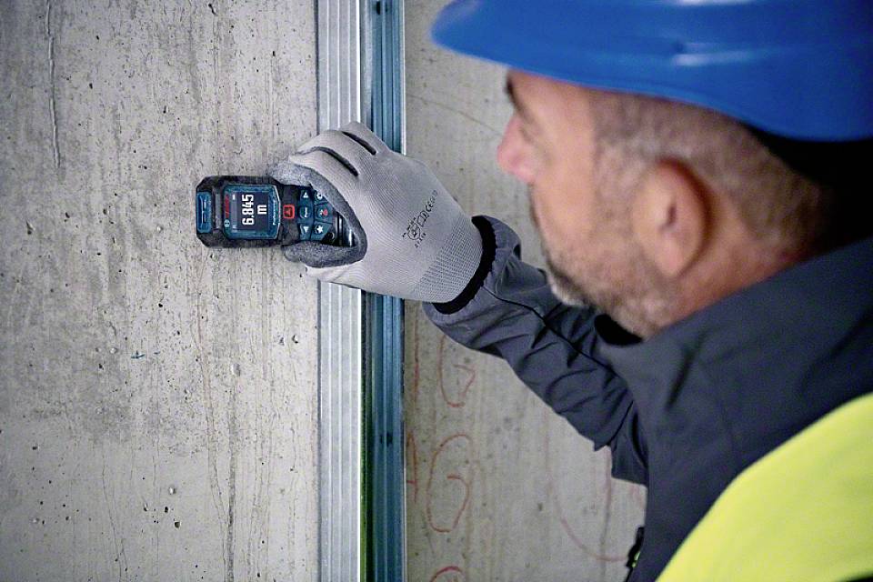 Dělník v modré přilce a rukavicích měří tloušťku stěny digitálním měřicím přístrojem na betonové zdi.