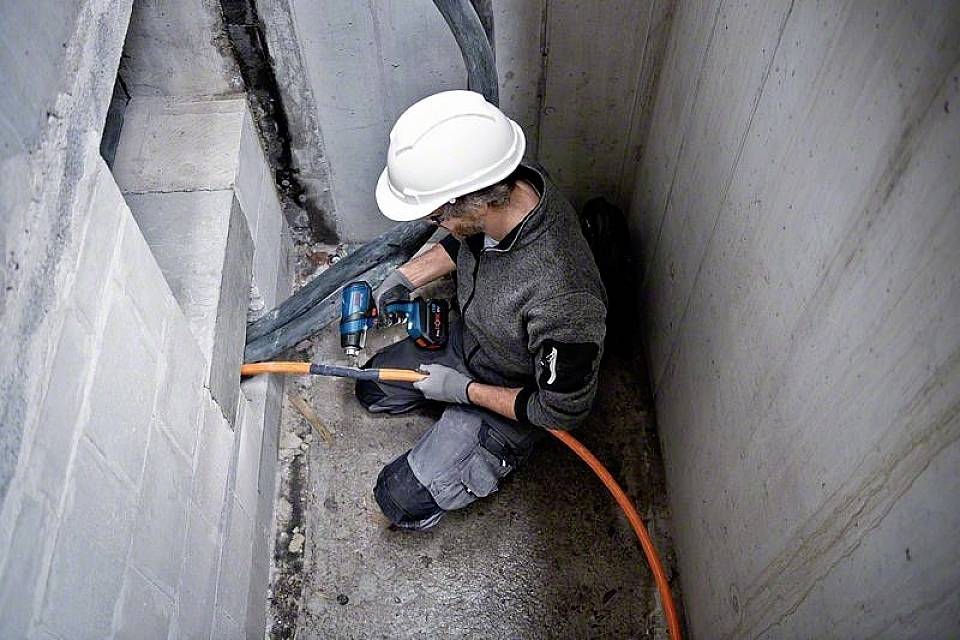 Dělník v helmě vrtá díru do betonové stěny a protahuje oranžový kabel. Nachází se v těsném prostoru.