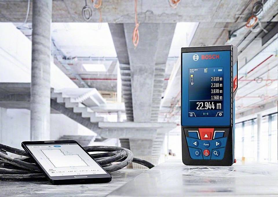 Digitální laserový dálkoměr na staveništi se smartphonem a diagramem aplikace pro měření délky, schodiště a kabely v pozadí.