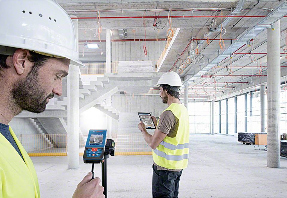 Stavební dělníci v ochranných oděvech používají digitální zařízení k měření v nedokončené budově. Moderní plánování staveb a technologie v akci.