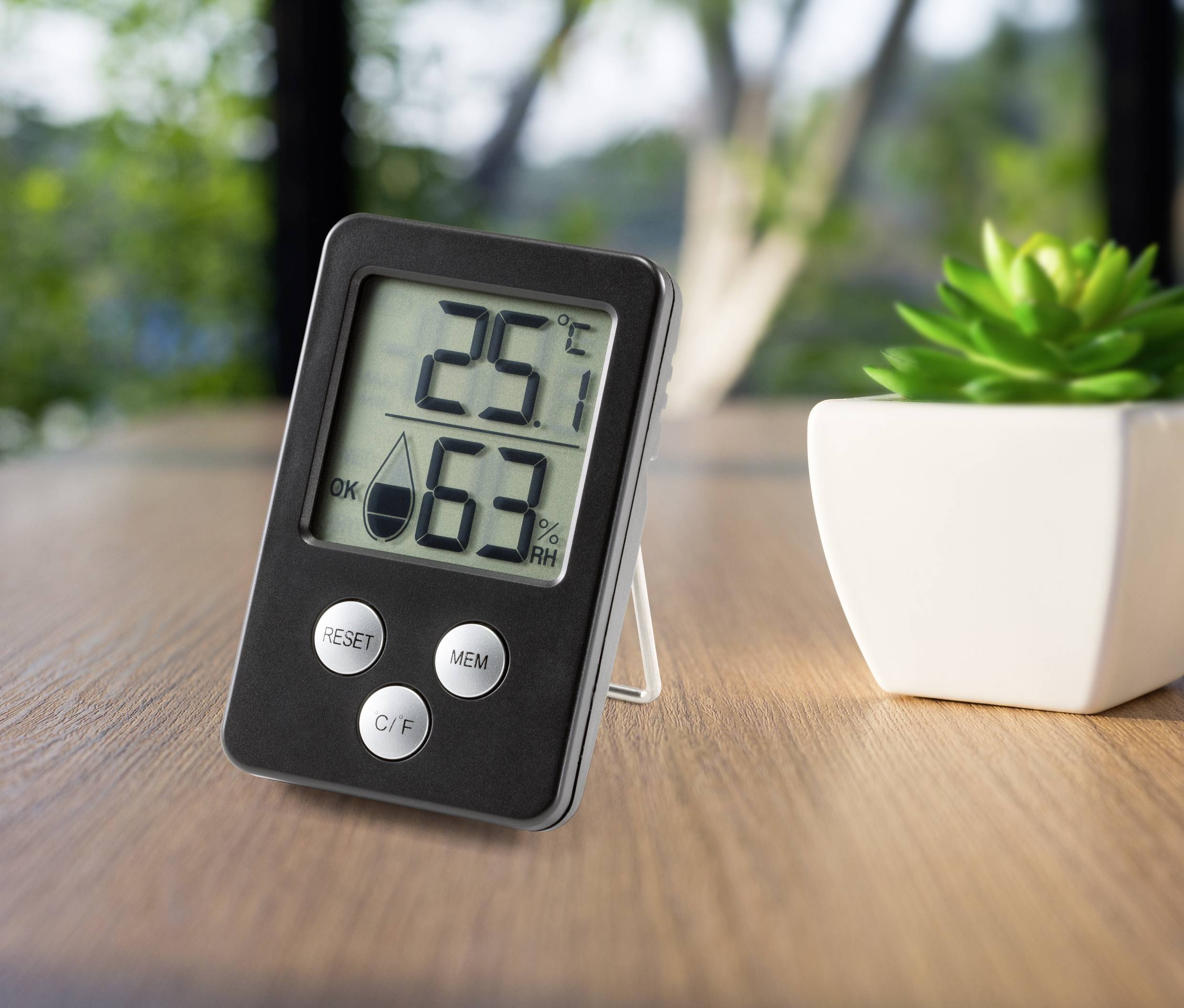 Digitální teploměr a vlhkoměr na stole ukazují 25°C a 63% relativní vlhkosti. V pozadí je rostlina.