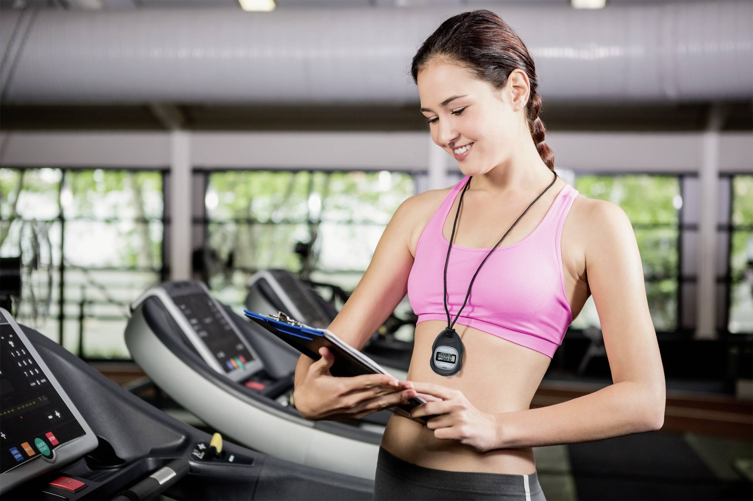 Žena ve fitness centru stojí s úsměvem před běžeckými pásy a drží poznámkový blok, zatímco kontroluje data na krokoměru.