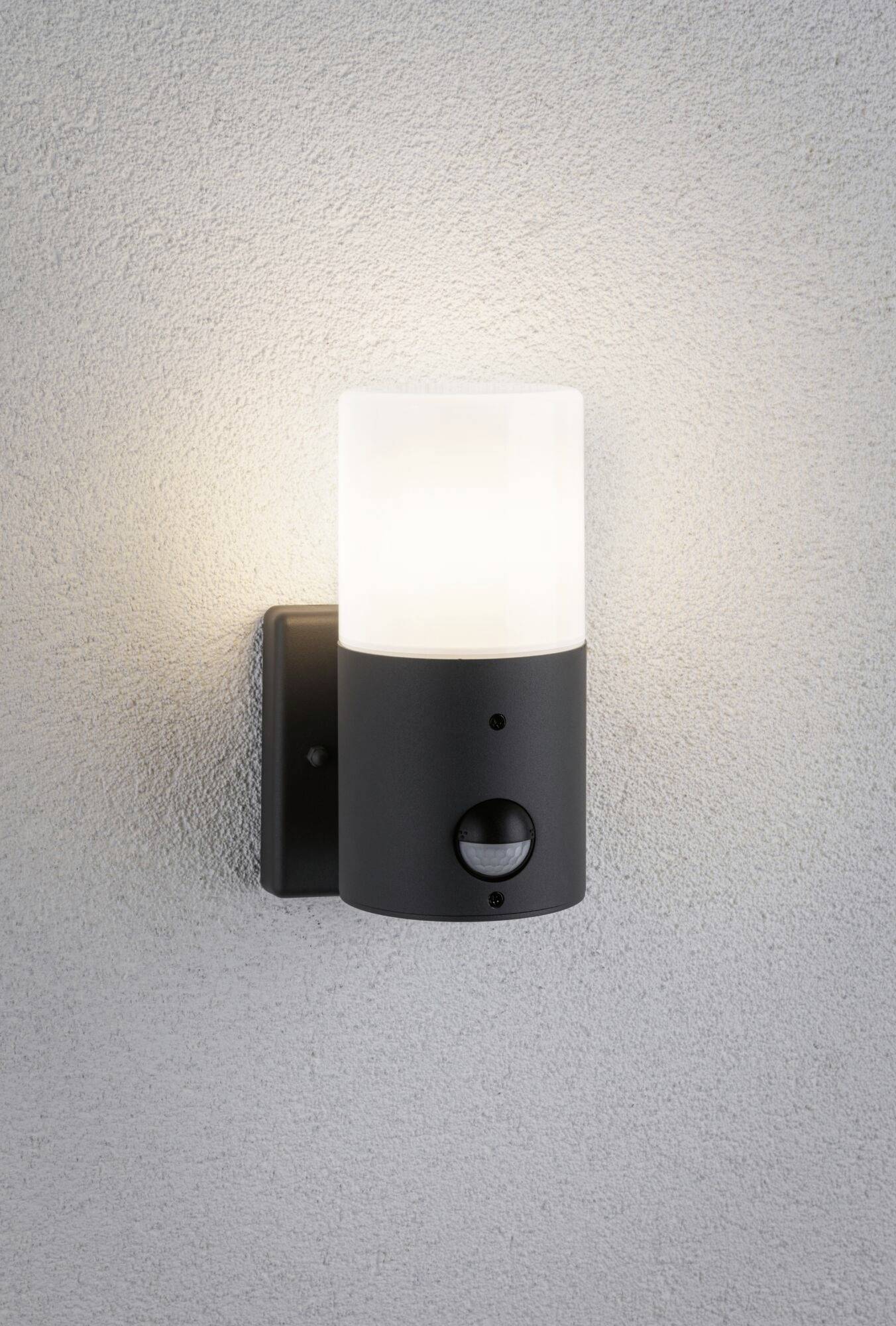 Venkovní lampa na stěně s černým tělem a bílým cylindrickým světelným difuzorem, pod nímž je umístěn pohybový senzor.