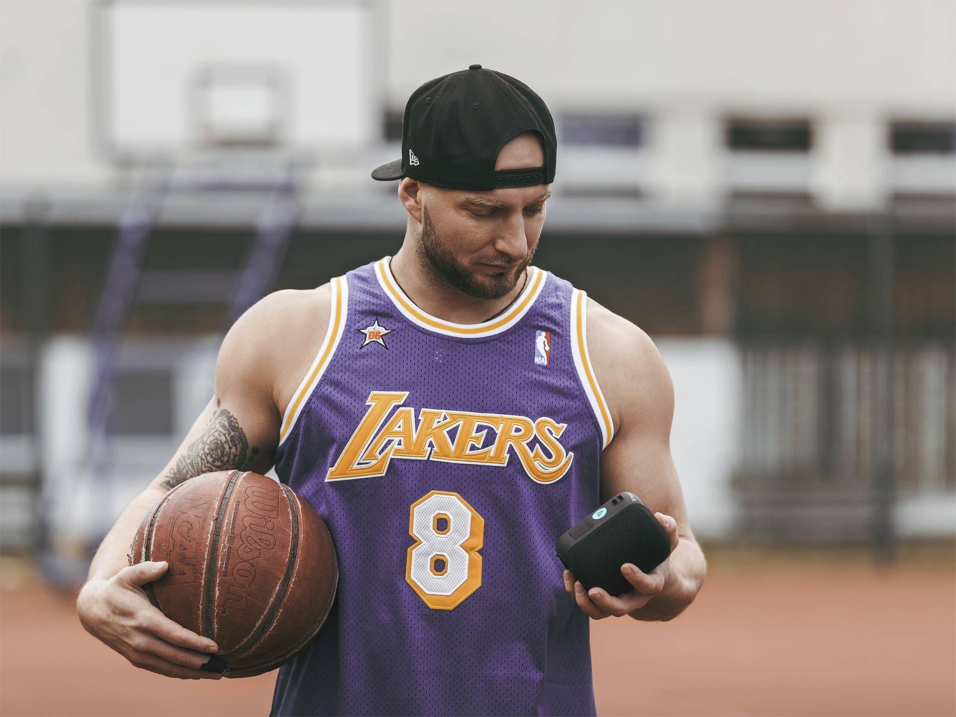 Muž v basketbalovém dresu s číslem 8 drží v jedné ruce basketbalový míč a v druhé malé zařízení. V pozadí se nachází basketbalové hřiště.