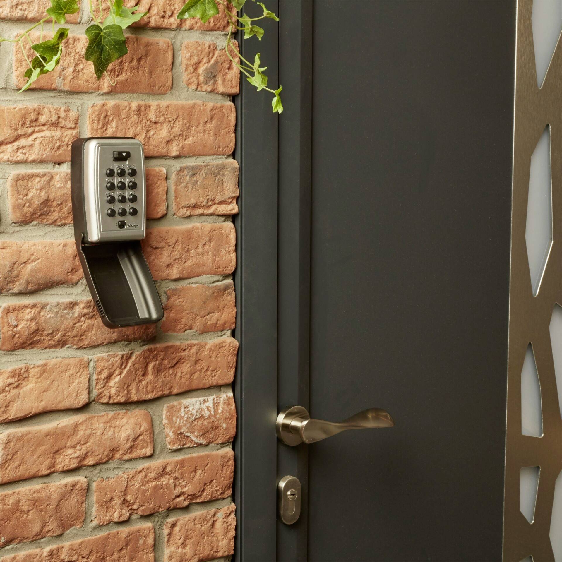 Nástěnný trezor s kombinačním zámkem vedle dveří na cihlové zdi, lehce otevřený, s visícími zelenými rostlinami.