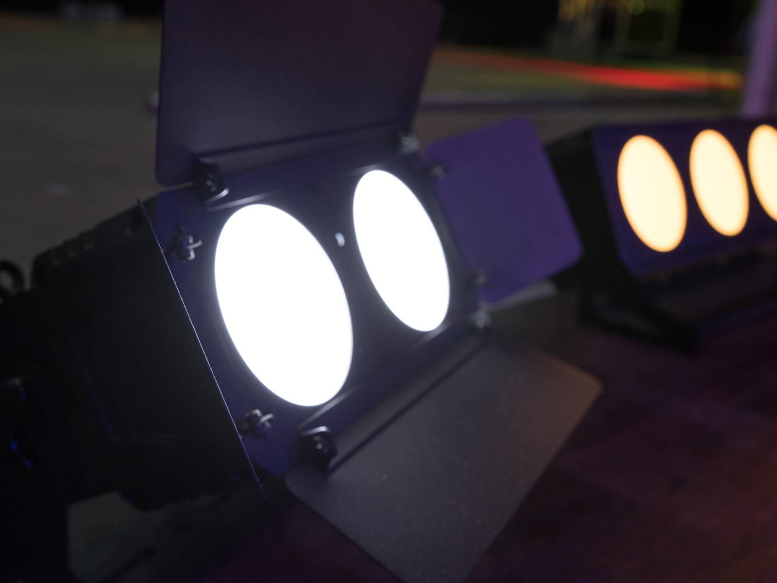 Jevištní reflektory se jasně rozsvítí, jeden bílým světlem, druhý oranžovým světlem. Jsou umístěny na dřevěné podlaze.