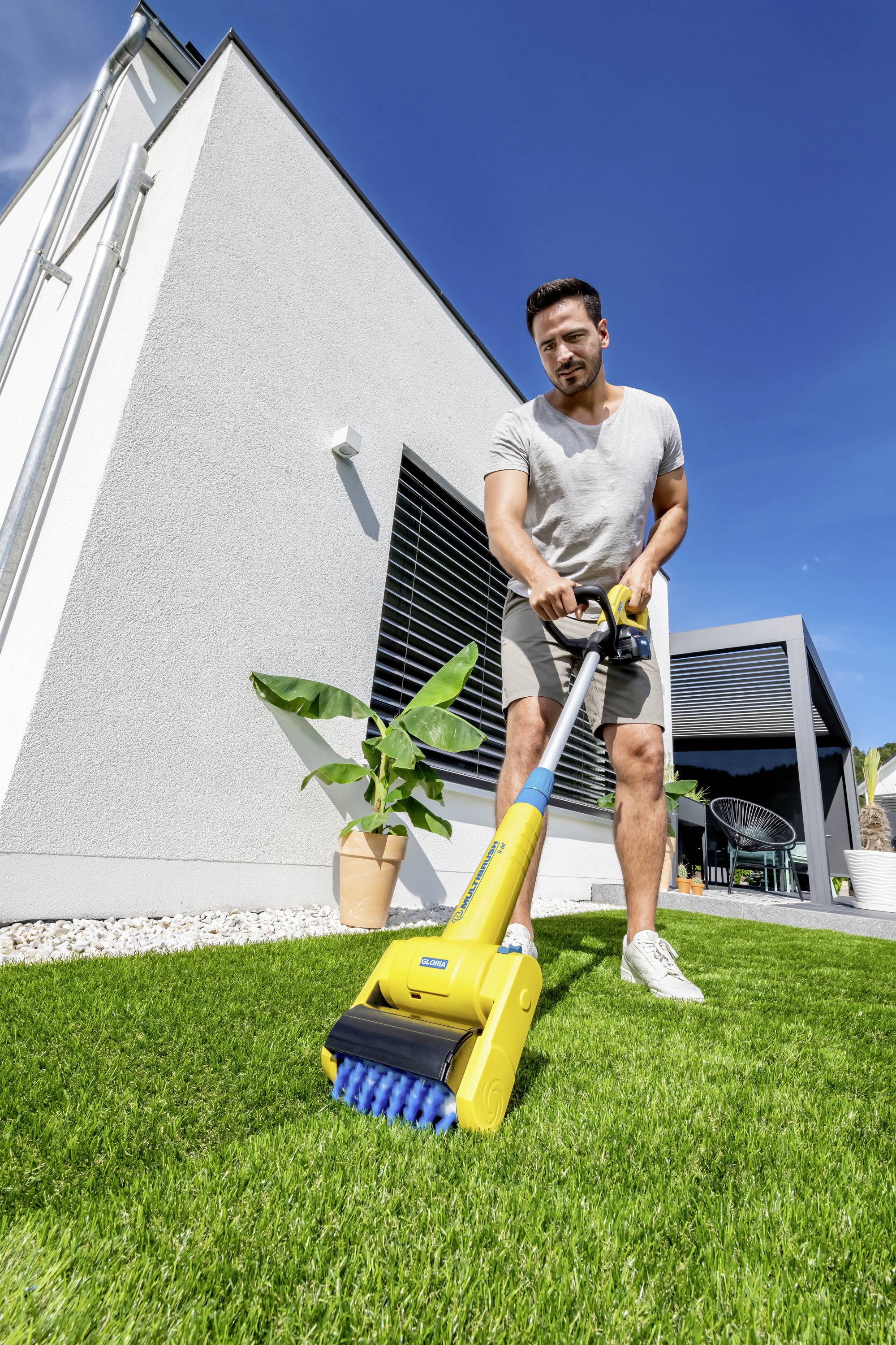 Muž udržuje trávník moderního domu pomocí žlutého čisticího zařízení. V pozadí jsou rostliny a modrá obloha.
