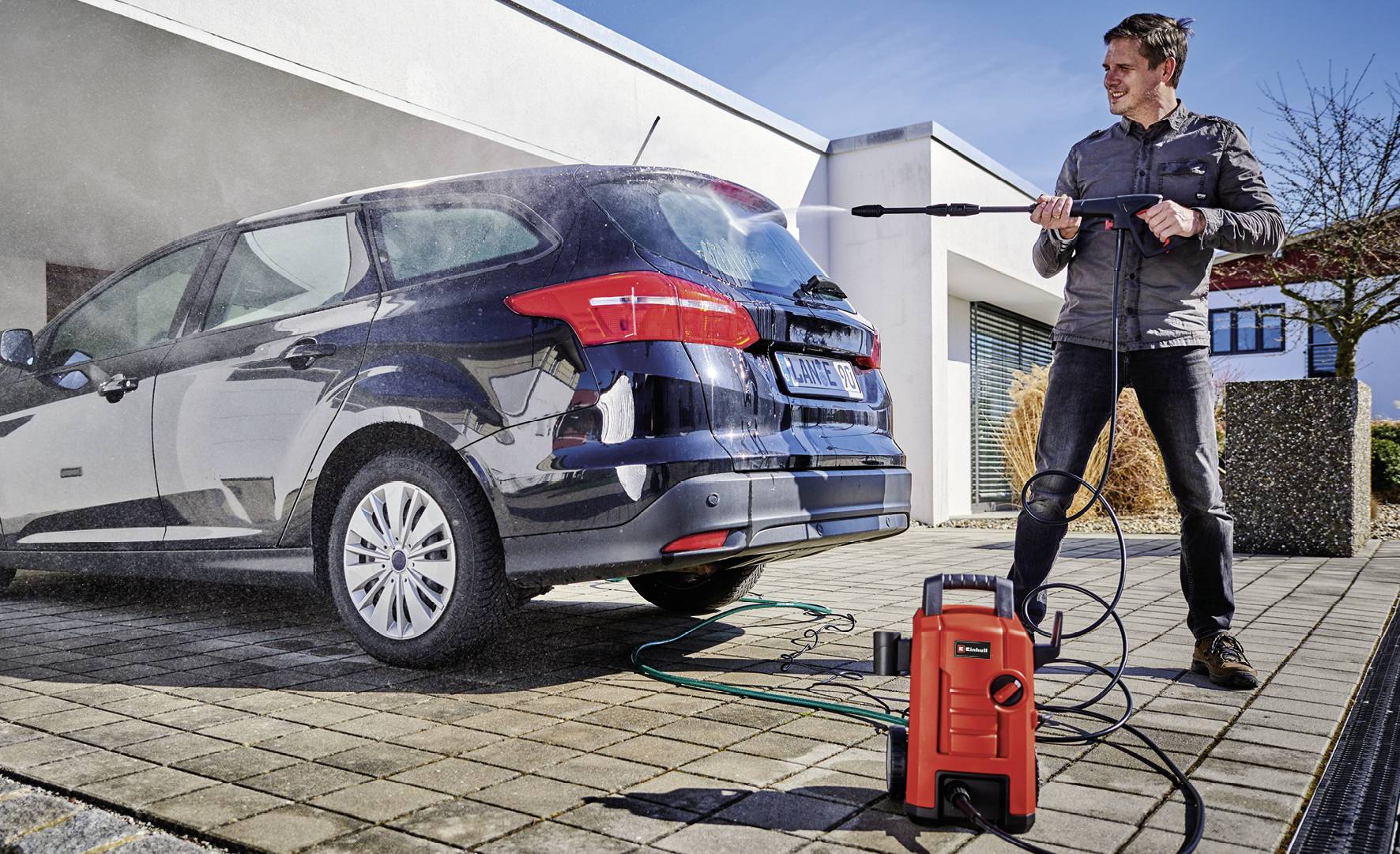 Osoba čistí černé auto vysokotlakým čističem na dlážděném vjezdu před moderním domem.