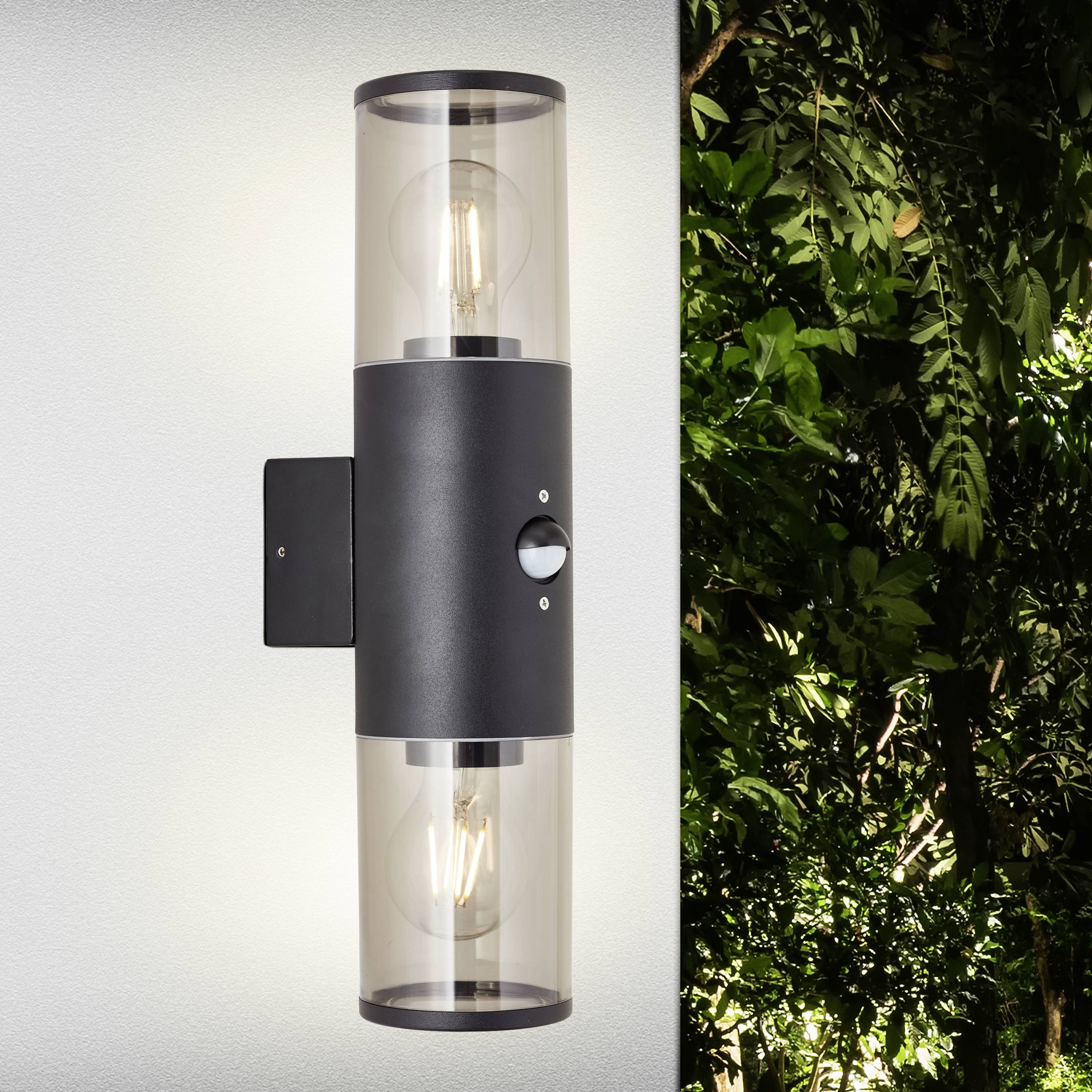 Nástěnná lampa s válcovitým dvojitým sklem na pozadí zeleného listí; ladí s přirozeným prostředím.