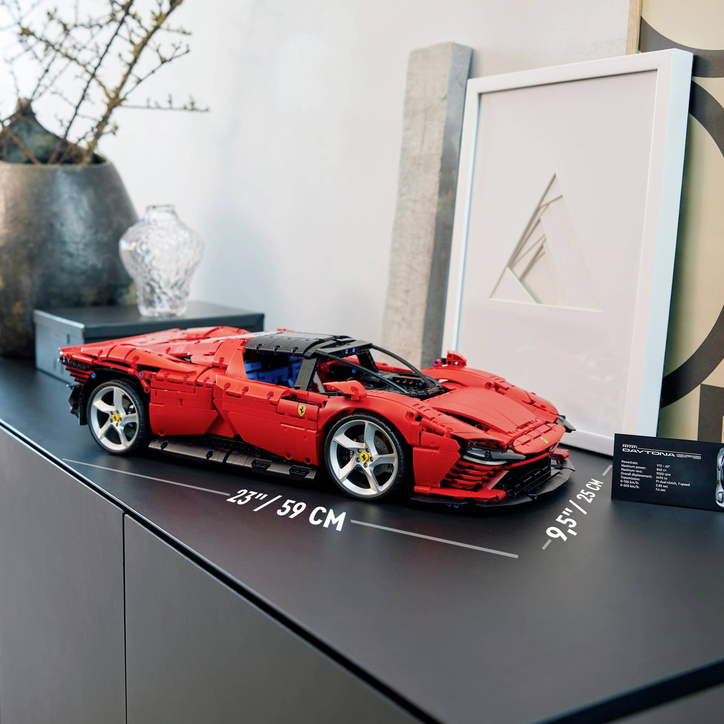 Červené modelové auto na stole vedle obrazu a rostliny. Měřítko ukazuje délku auta 59 cm.